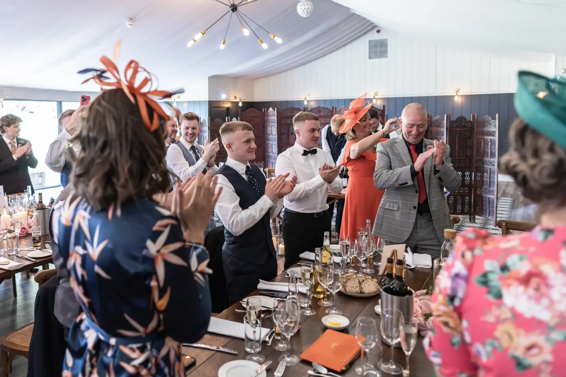 Guests clapping for a couple at a wedding reception inside a tent, with some attendees in elegant hats.