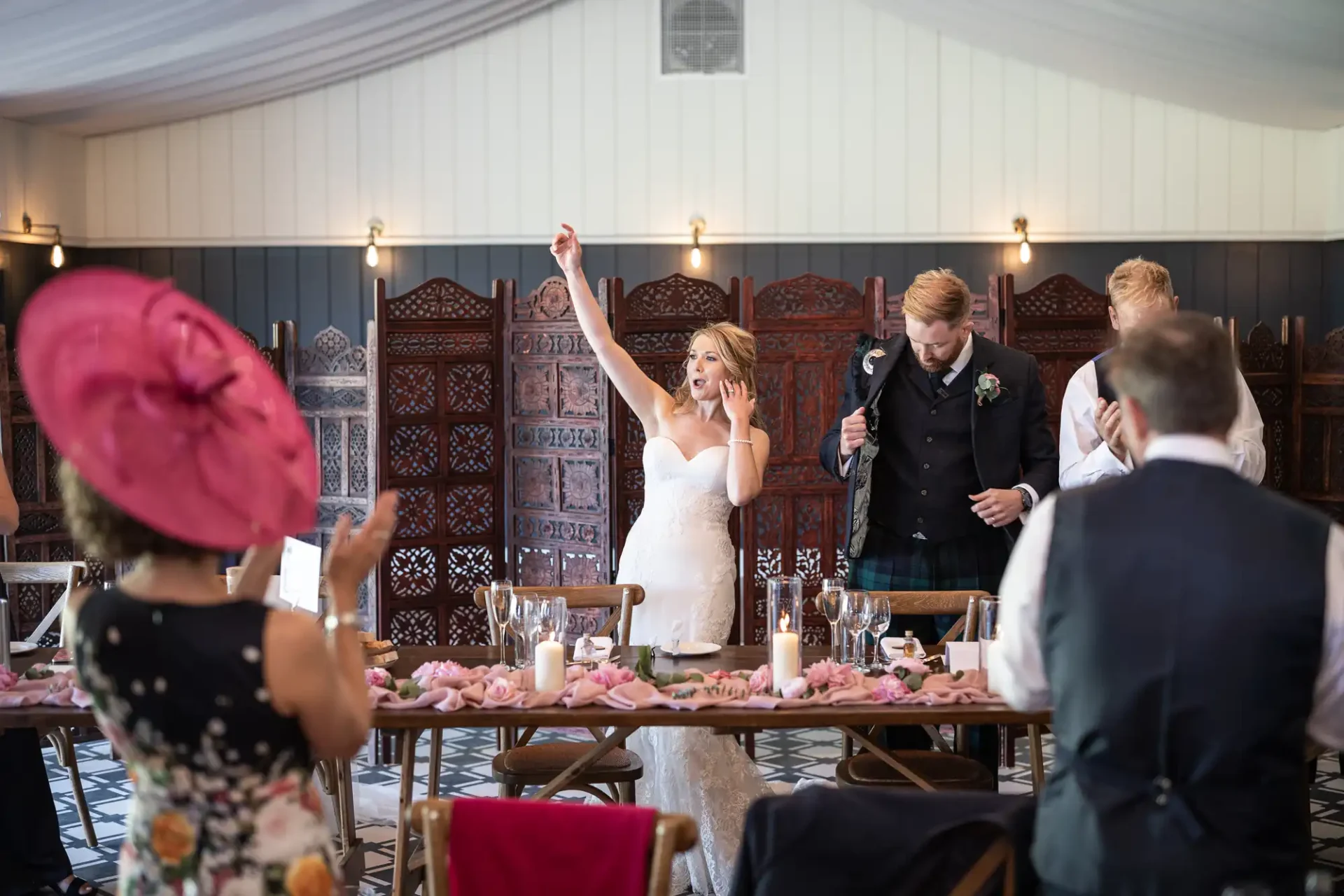 A bride joyfully raises her arm at a wedding reception as guests look on, surrounded by elegantly decorated tables.