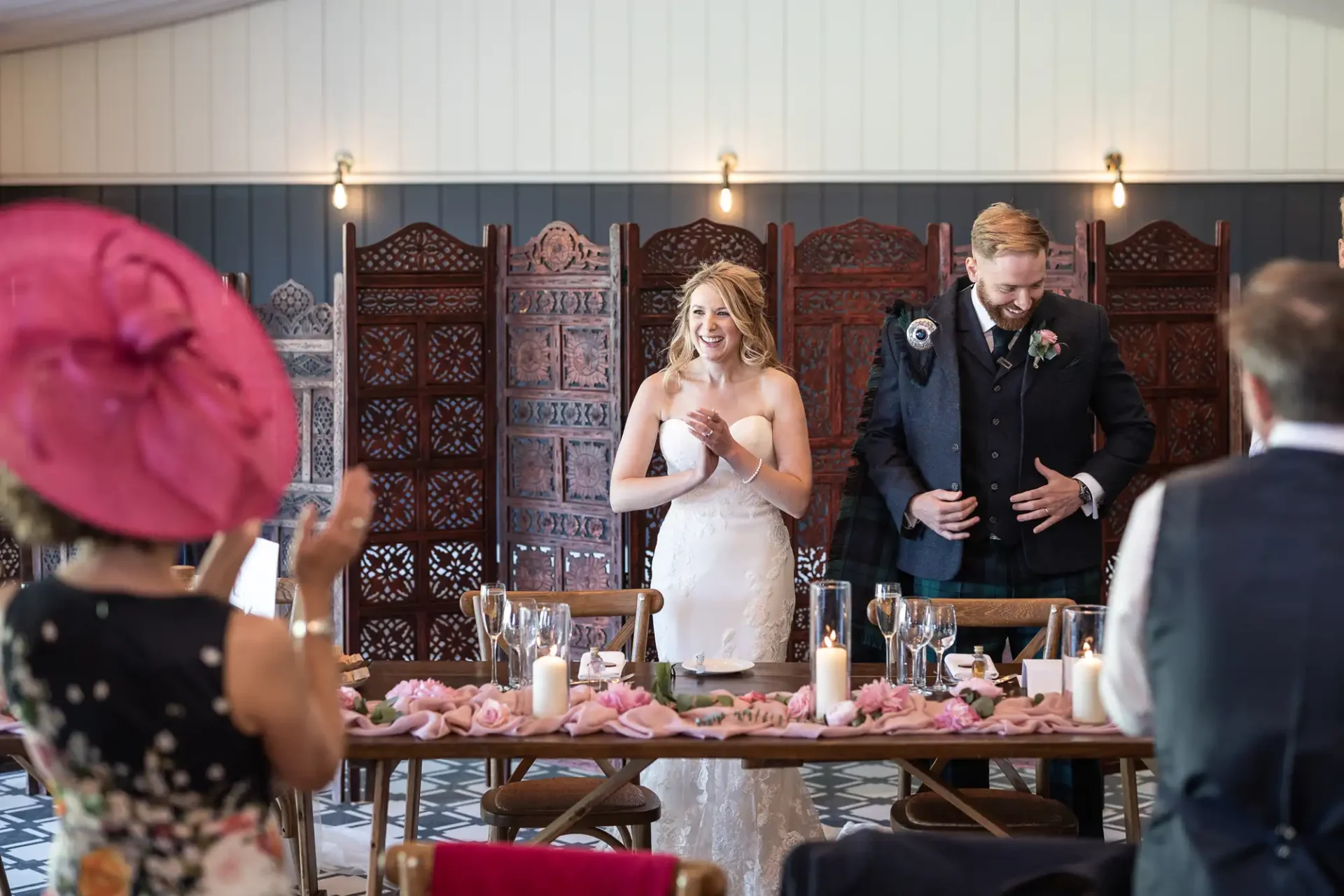 Bride and groom smiling at a wedding reception while guests look on, groom wearing a kilt, beautifully decorated table with pink flowers.