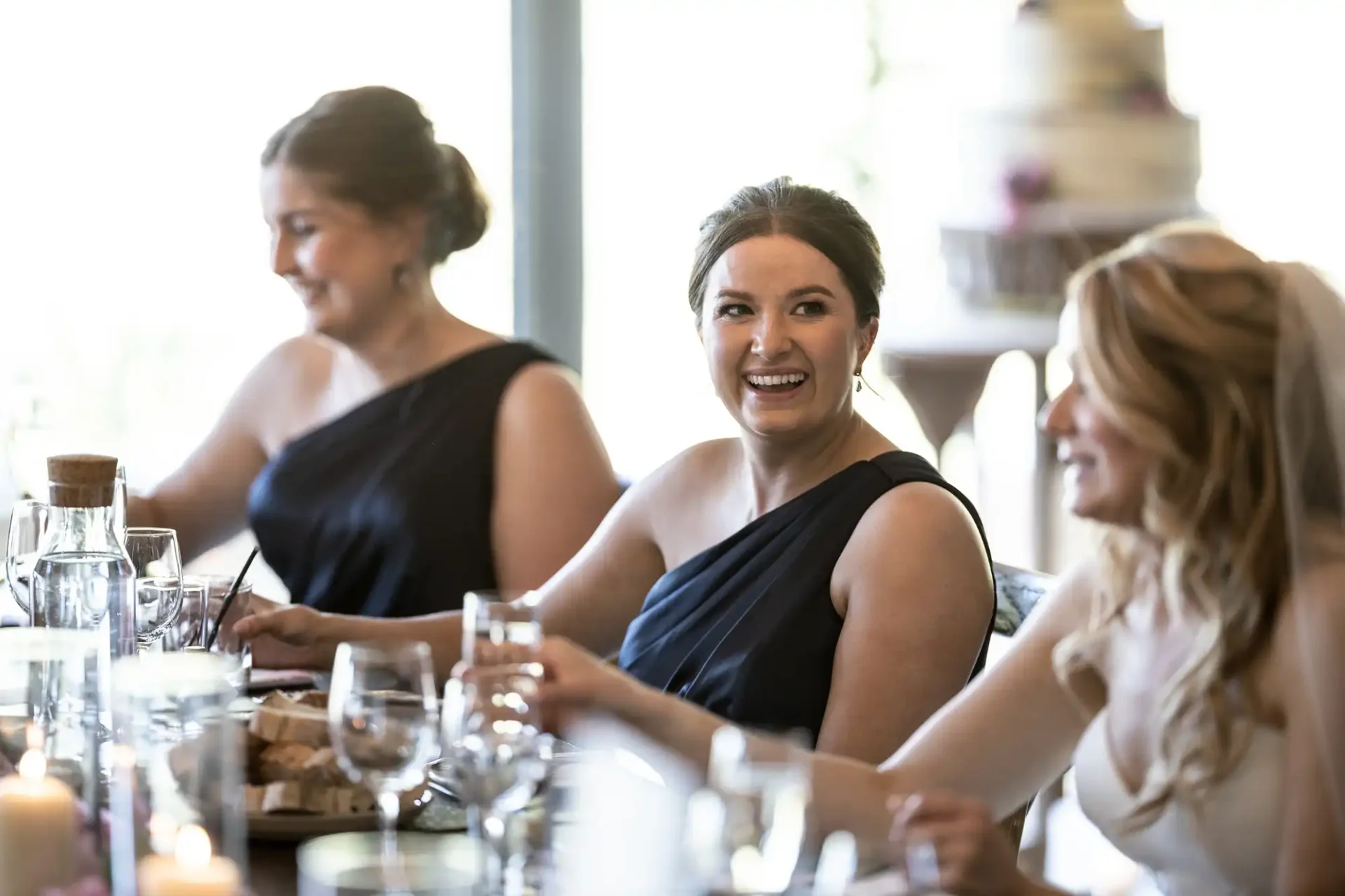 Three women in elegant black dresses smiling and sitting at a dining table during a festive event.
