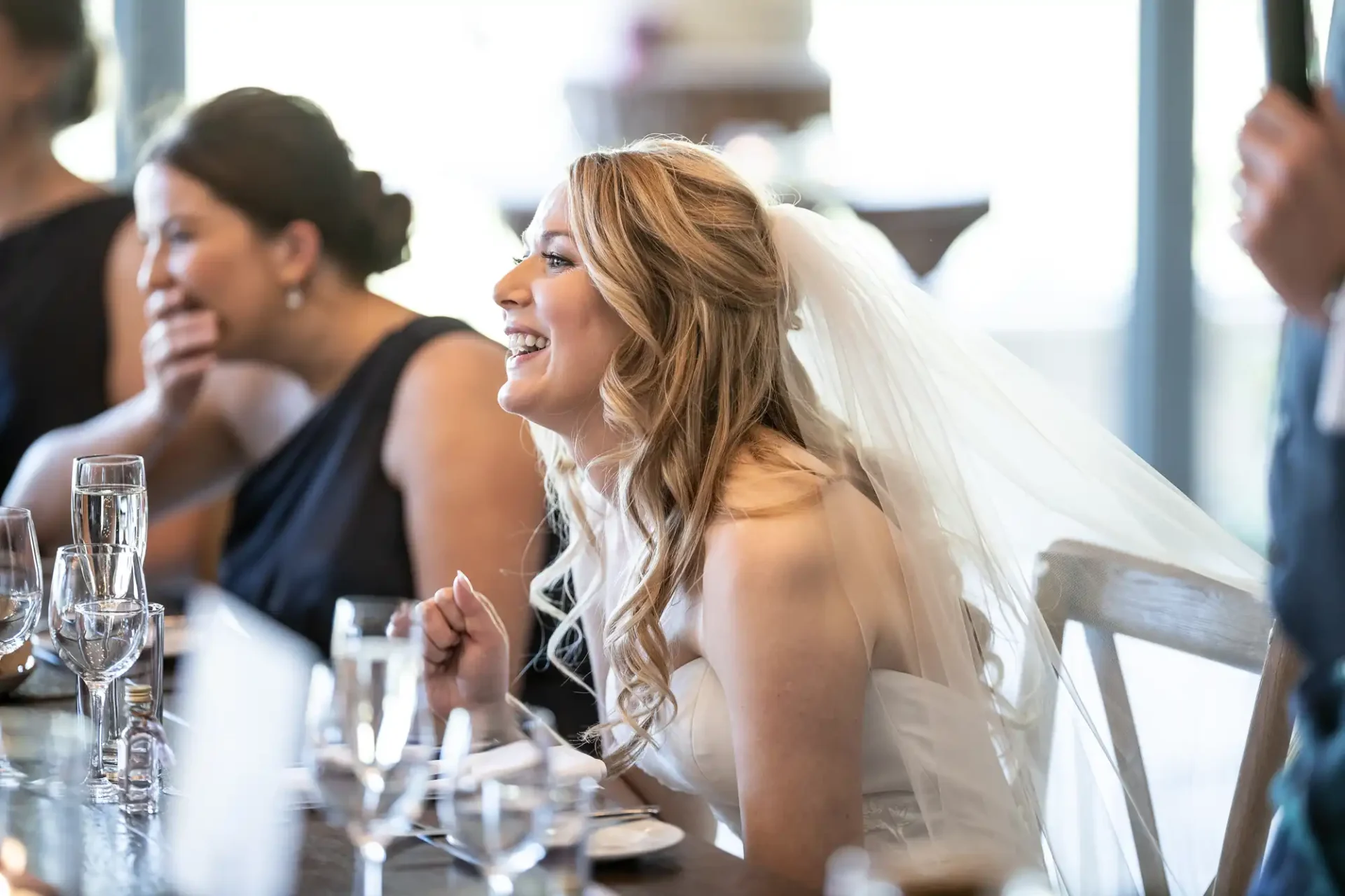 A bride in a white dress with a veil laughs joyfully at a table during a wedding reception, surrounded by guests.