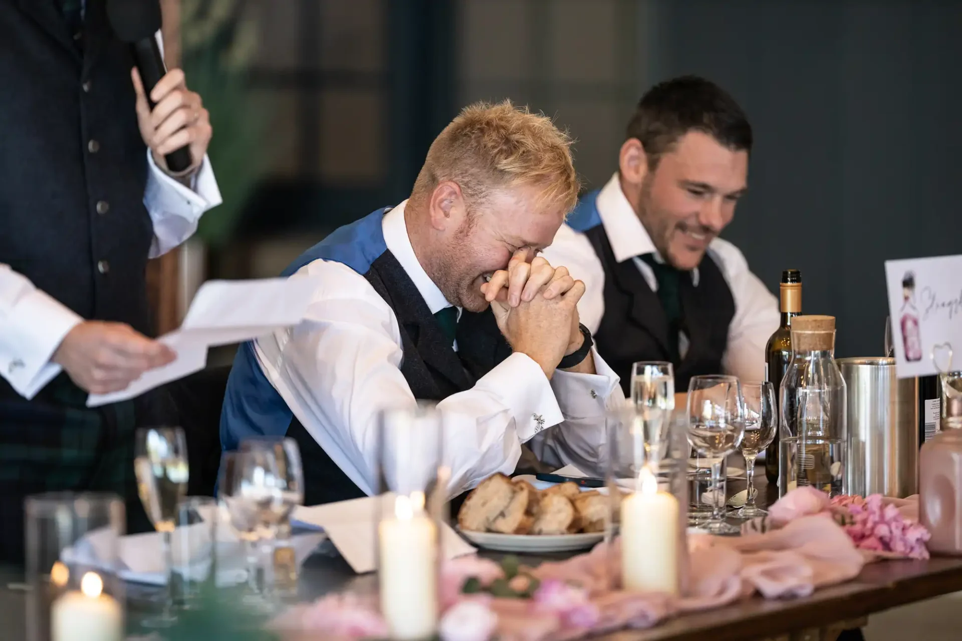 A man in a vest laughs with his face in his hands at a dining table during a speech, with another man smiling beside him.
