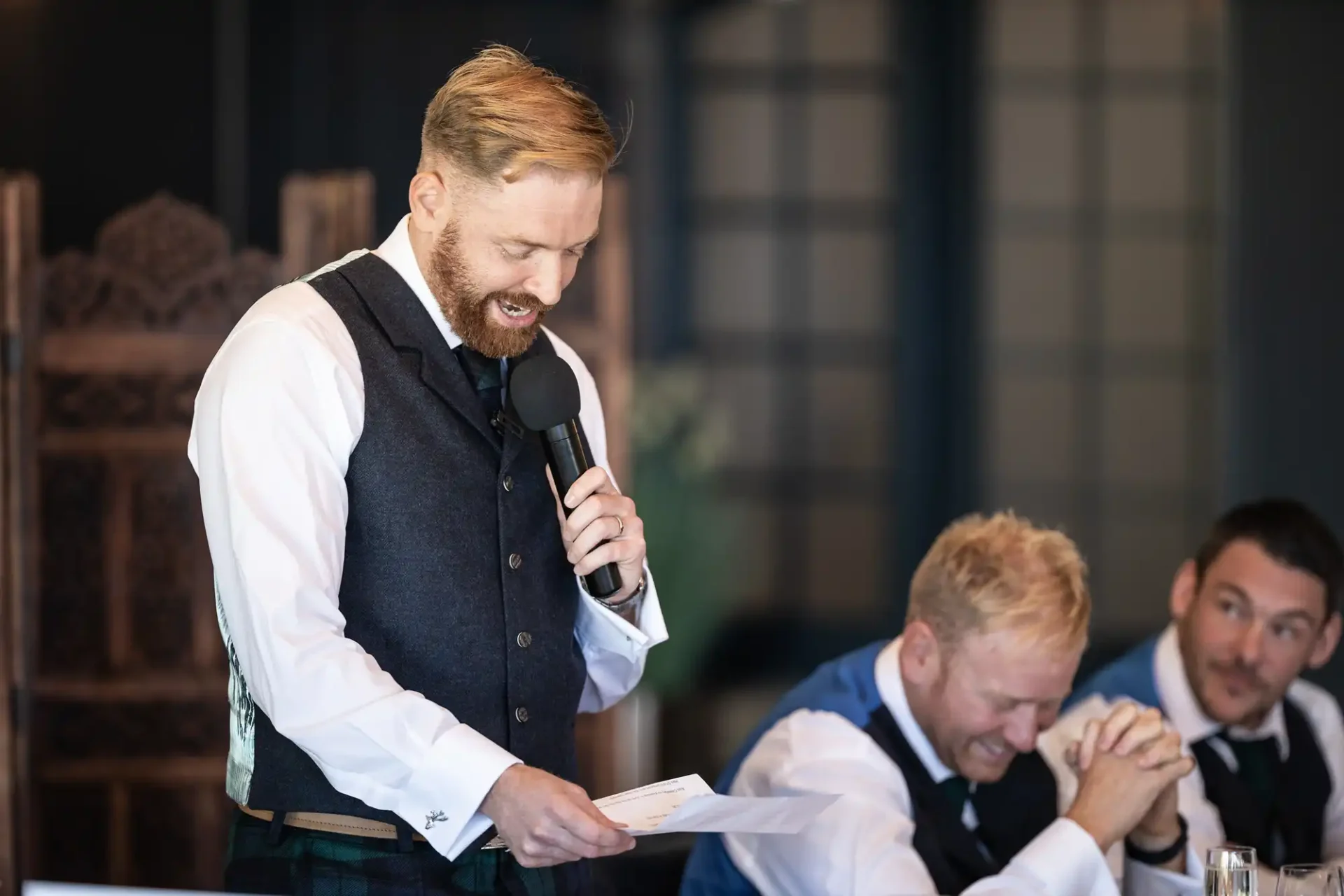 A man with a beard, wearing a vest and kilt, gives a speech holding a microphone and a paper, with another man laughing in the background.