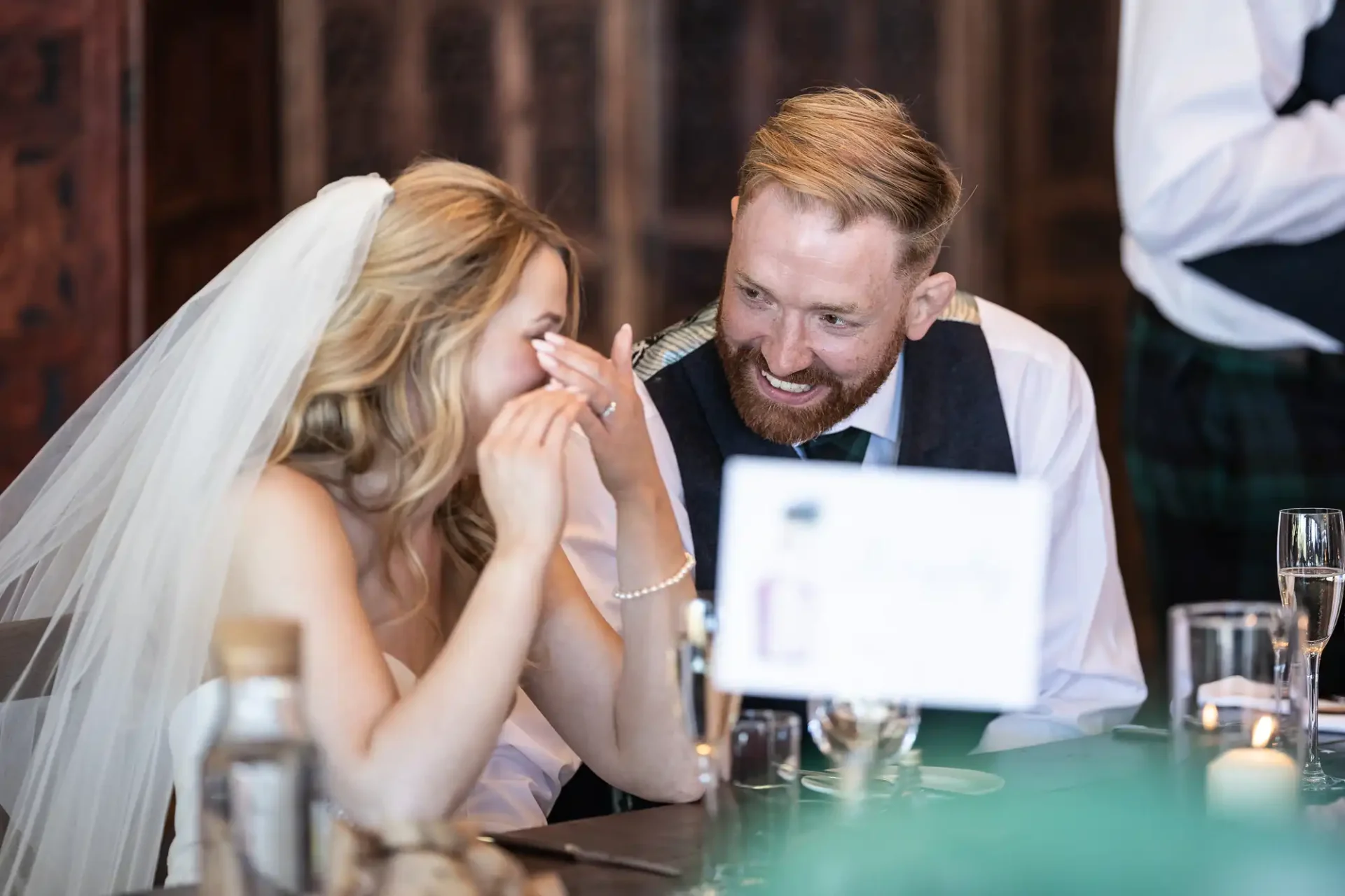 Bride and groom laughing together at a wedding reception table, with the groom wearing a suit and the bride in her wedding dress.