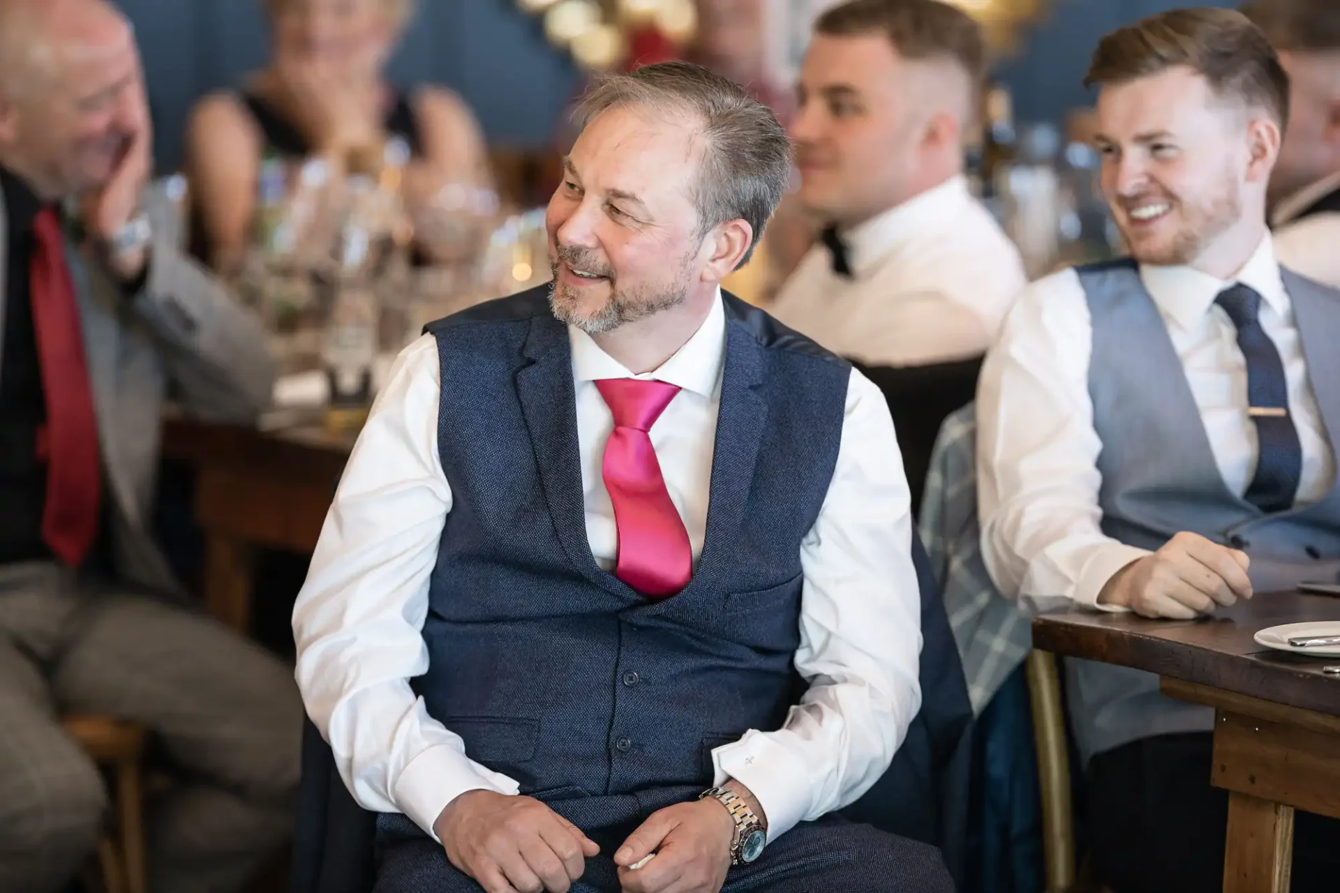 A man in a vest and red tie smiles while sitting at a festive event, surrounded by other guests also in semi-formal attire.