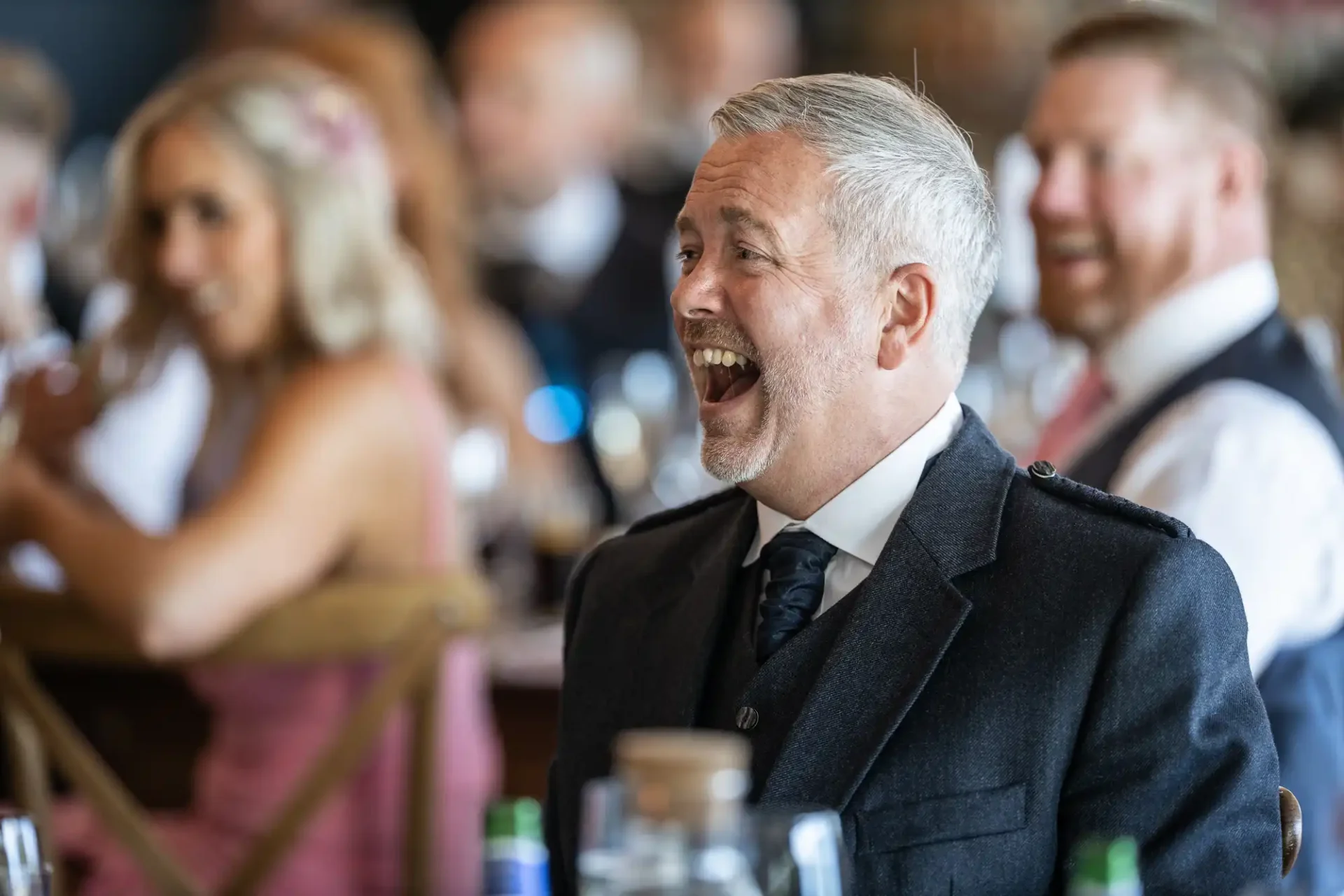 A middle-aged man with gray hair, wearing a suit and tie, laughing joyously at a social event with people in background.