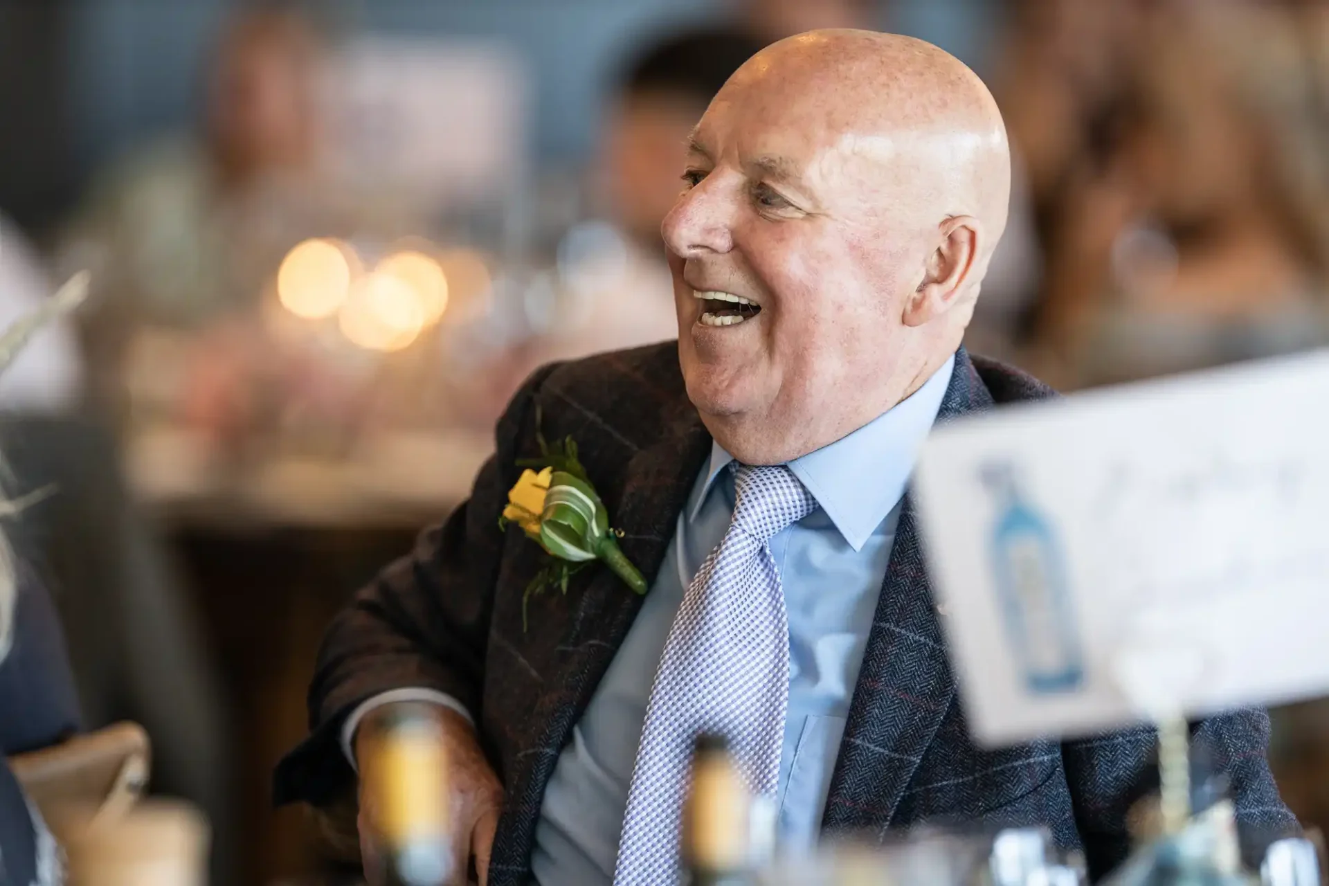 Elderly man in a suit with a boutonniere, laughing at a social event with other guests blurred in the background.
