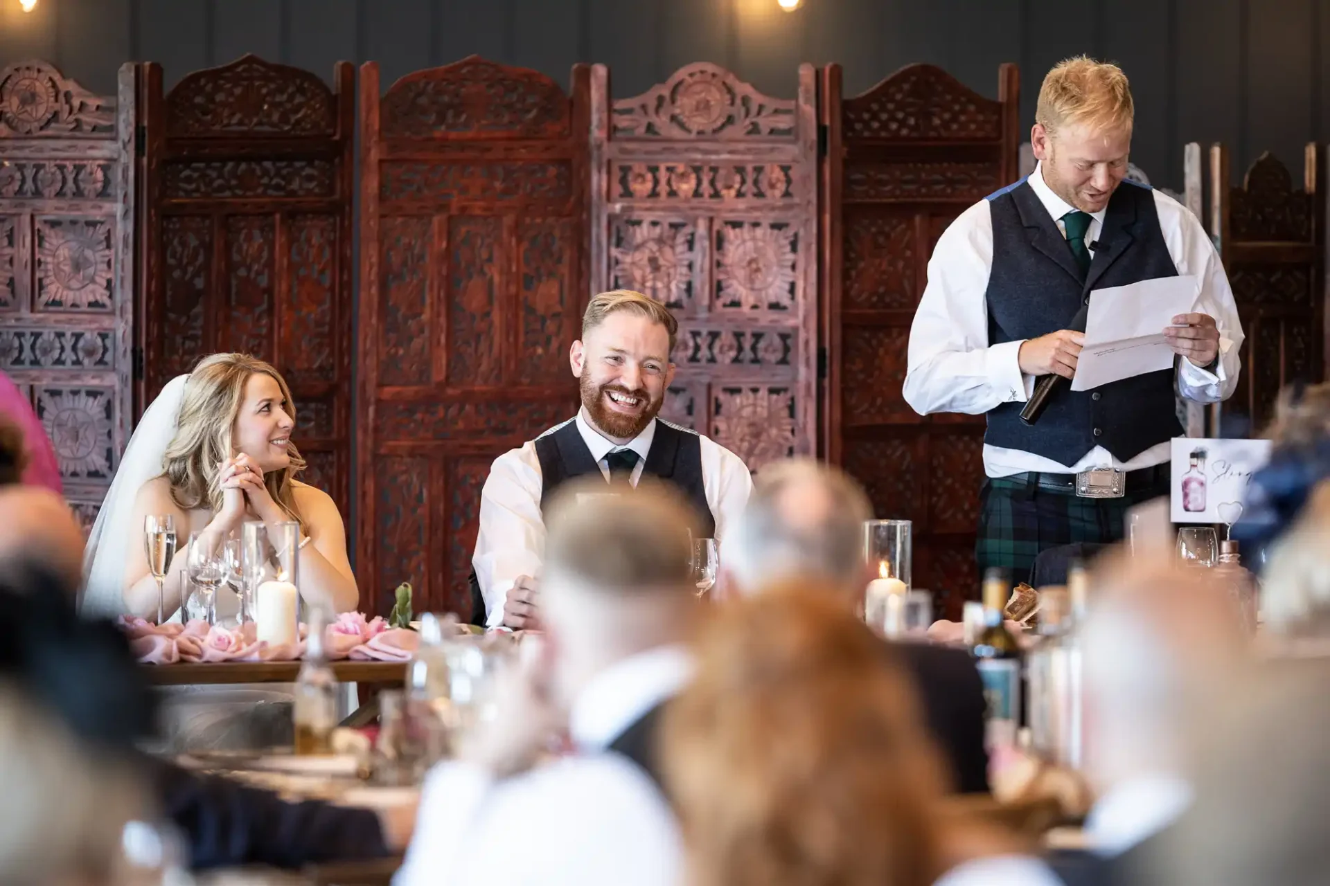A man in a kilt delivers a speech at a wedding, with a smiling bride and groom seated at a table, surrounded by guests in a banquet hall.