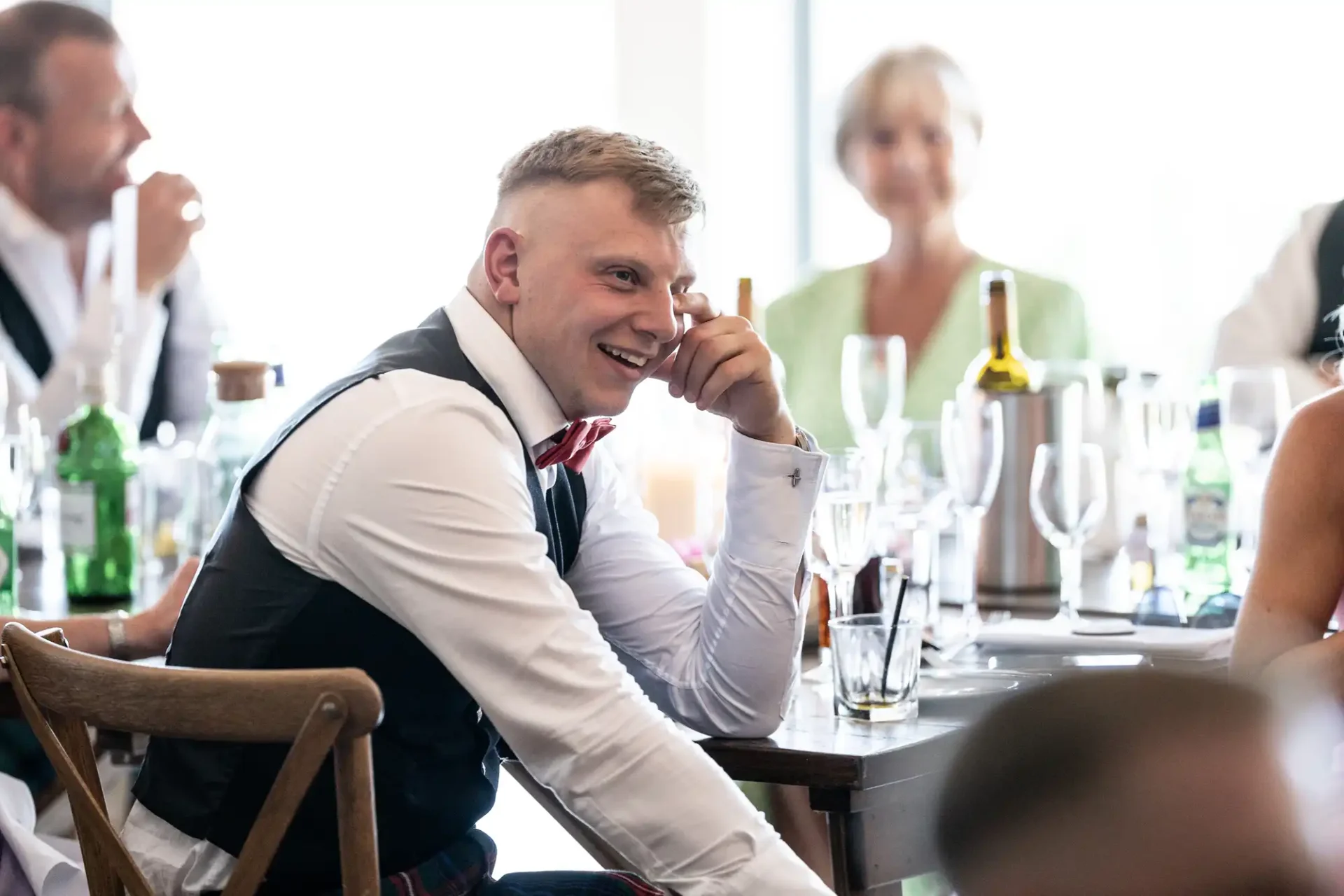 Man in a formal vest and tie smiling and talking on a cellphone at a busy wedding reception table.