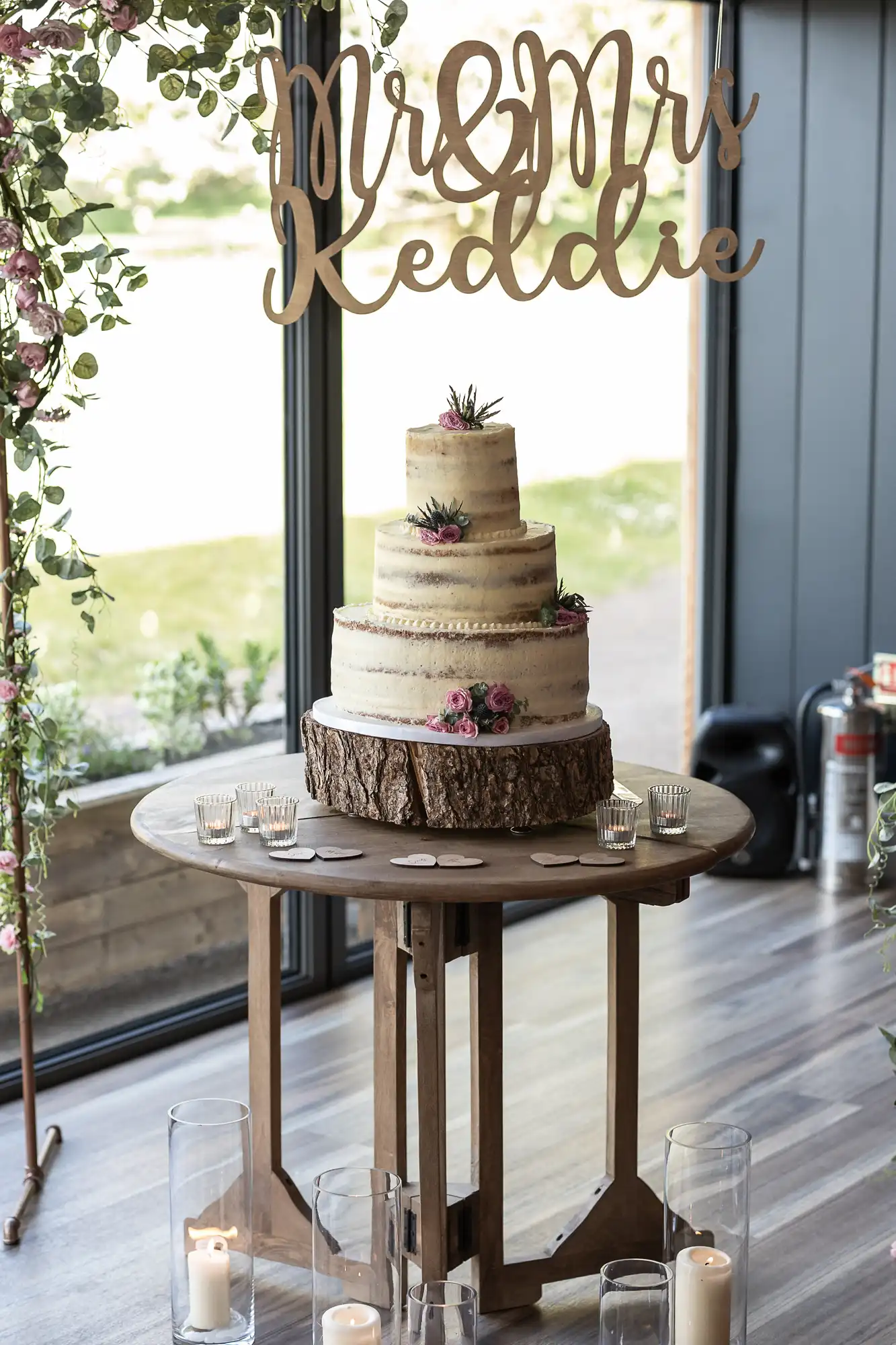 A three-tiered wedding cake decorated with floral accents on a wooden stand, displayed under a "mr & mrs" sign by a window, surrounded by lit candles.