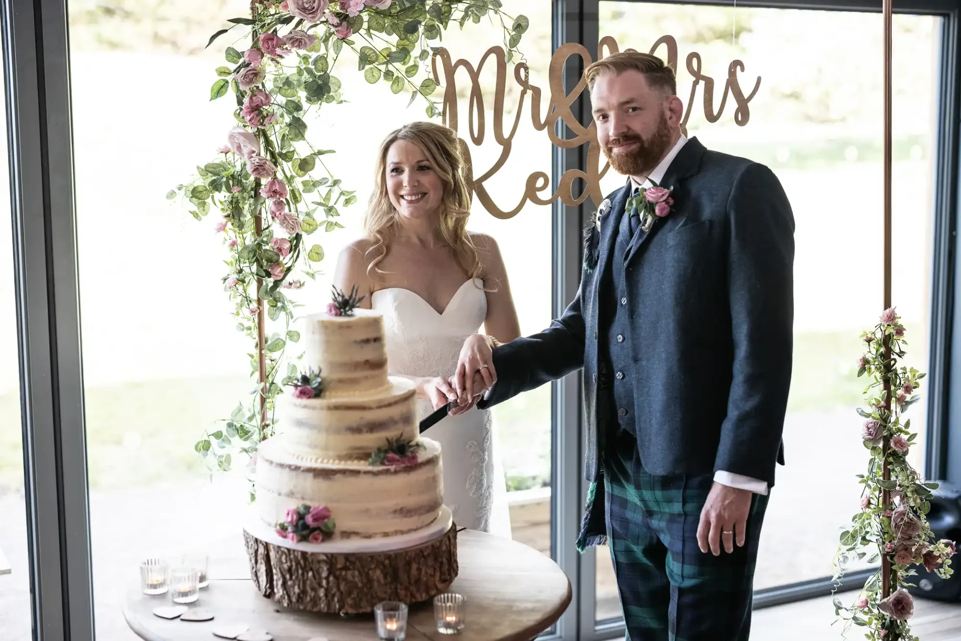 A bride and groom happily cutting a three-tier wedding cake, standing by a floral archway with a "mr & mrs" sign above.