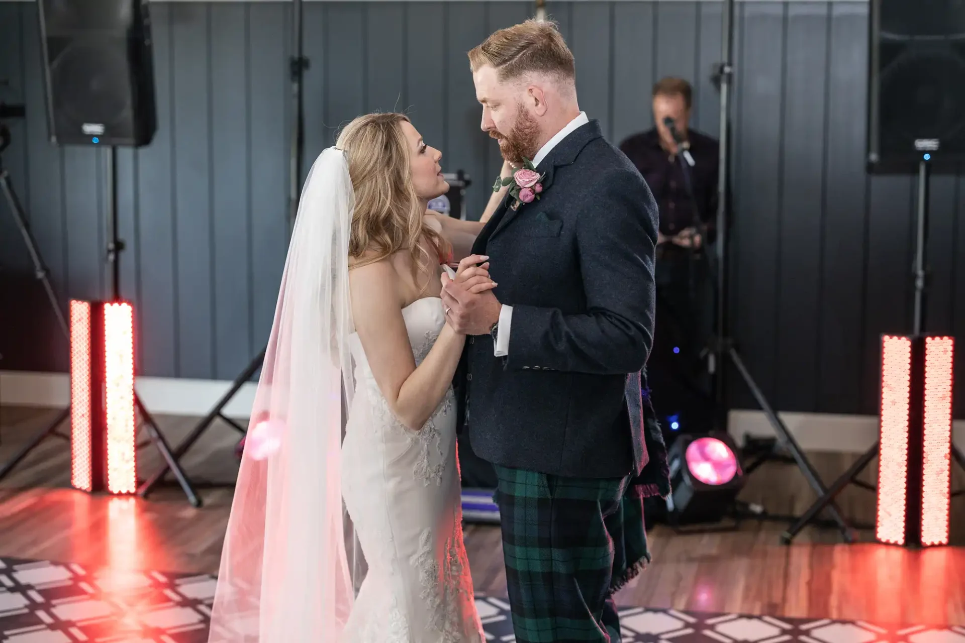 A newlywed couple dances closely at their wedding reception, with the bride in a white gown and the groom in a kilt, against a backdrop of colorful lights and a live band.