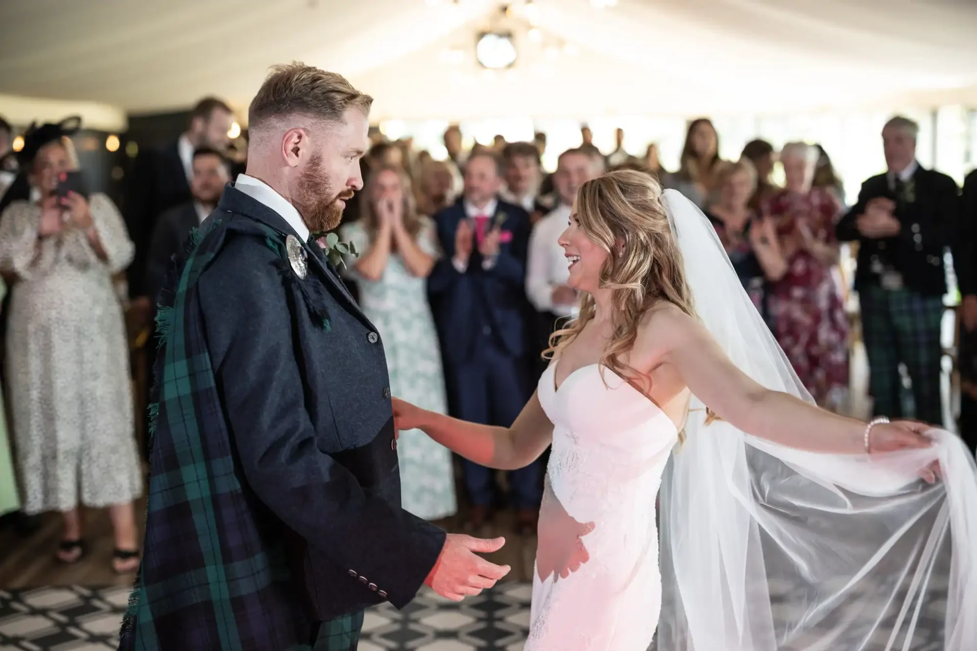 A bride in a white dress and a groom in a tartan kilt hold hands, smiling joyously, during a wedding ceremony with guests in the background.