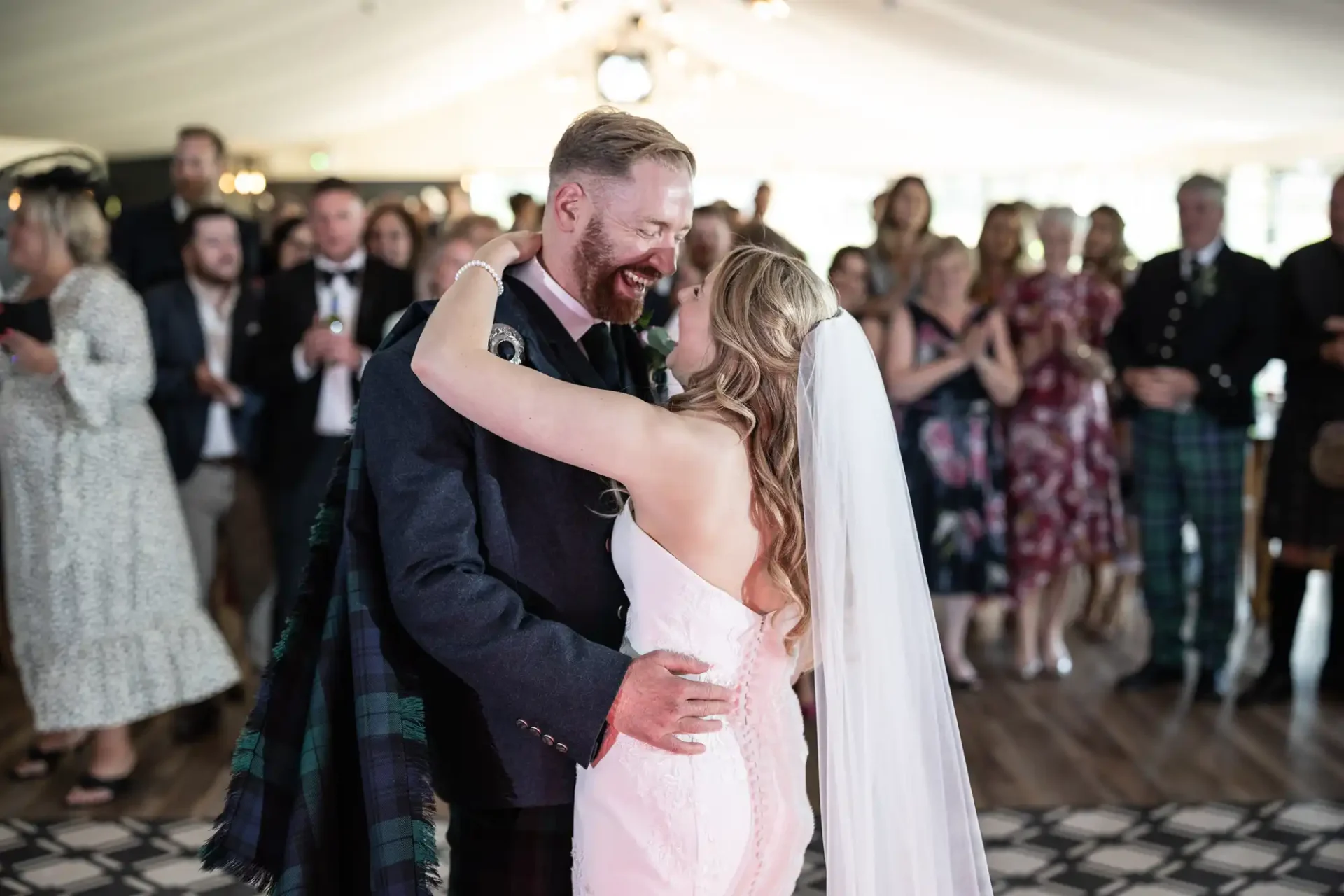 Bride and groom joyfully embracing during their wedding dance in a tent, surrounded by guests.