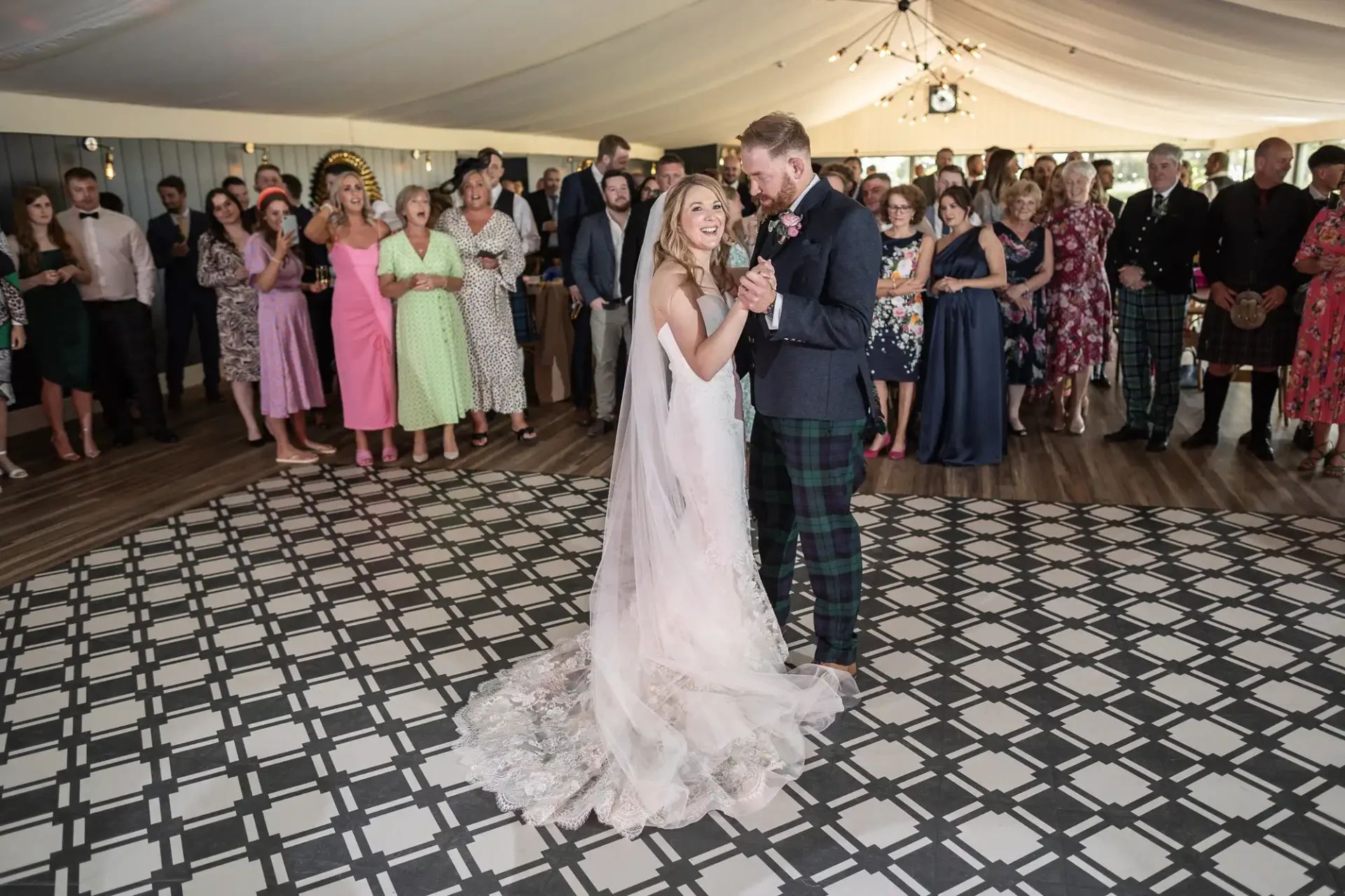 A bride and groom share their first dance surrounded by guests in a tent with a checkered floor.