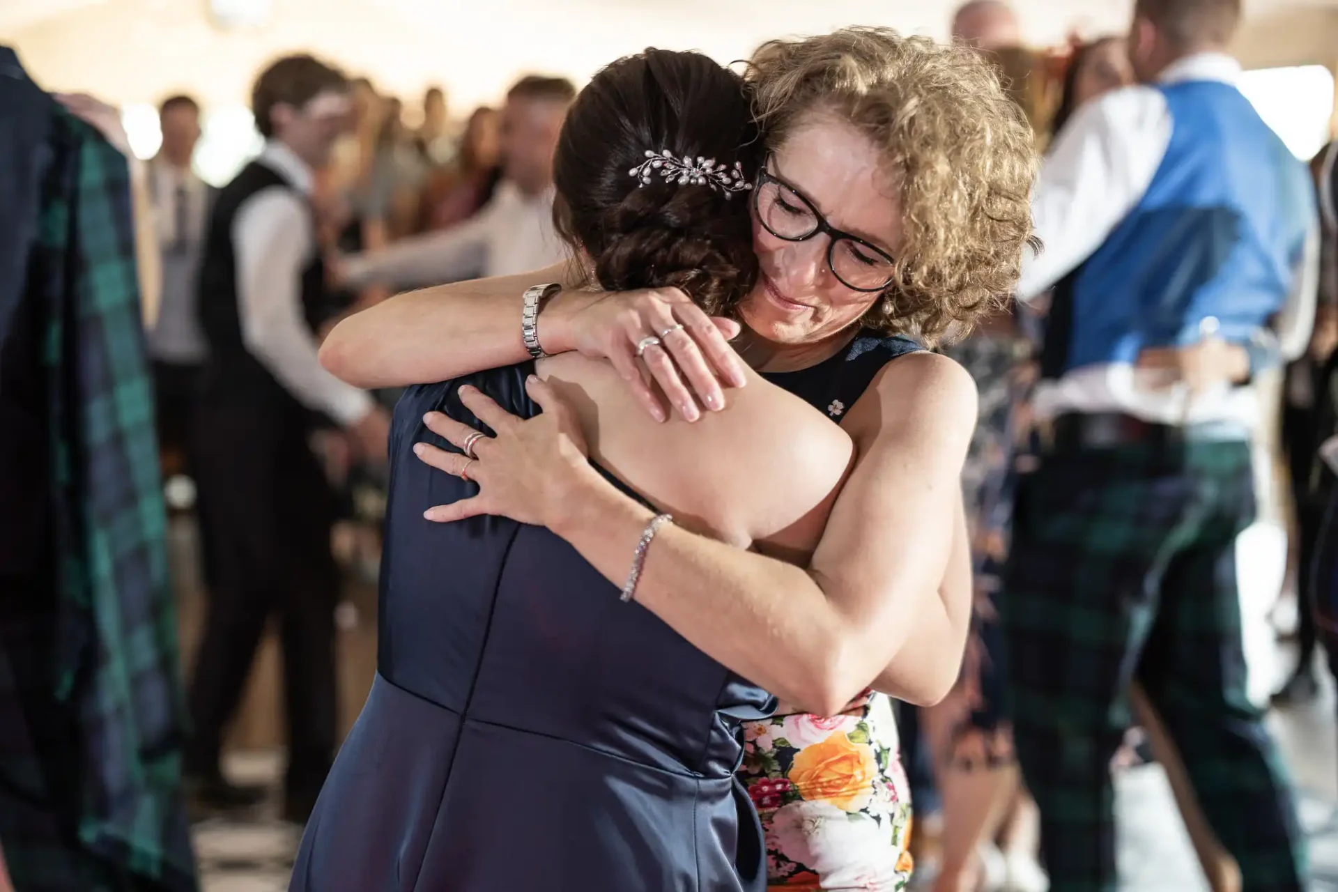 Two women embracing warmly at a dance event, surrounded by other attendees, one wearing a floral dress and the other in a navy dress with glasses.
