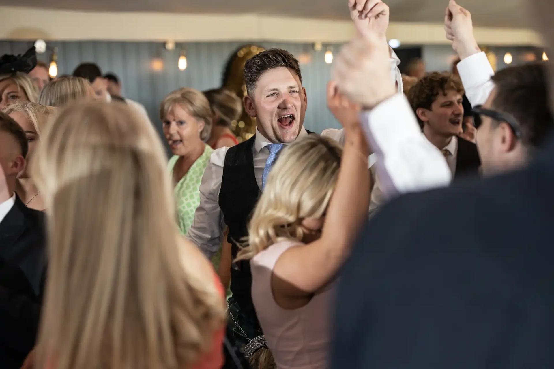 A joyful man raises his arm among a crowd of dancing people at an indoor celebration.