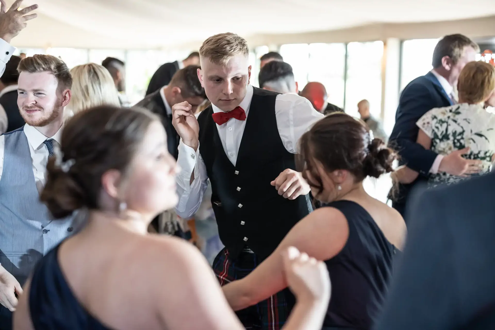 Young man in a vest and kilt dancing with a woman in a blue dress at a lively wedding reception.