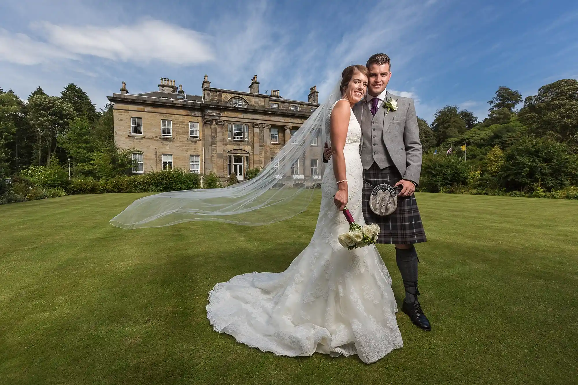 A bride in a white dress and a groom in a kilt smiling on a grass lawn with a stately home in the background on a sunny day.
