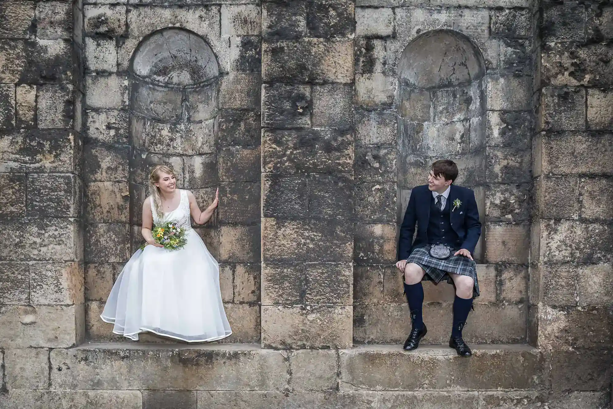 Bride in a white dress and groom in a kilt sitting apart on ledges against an old stone wall, looking at each other - Wedding-photography prices in Scotland