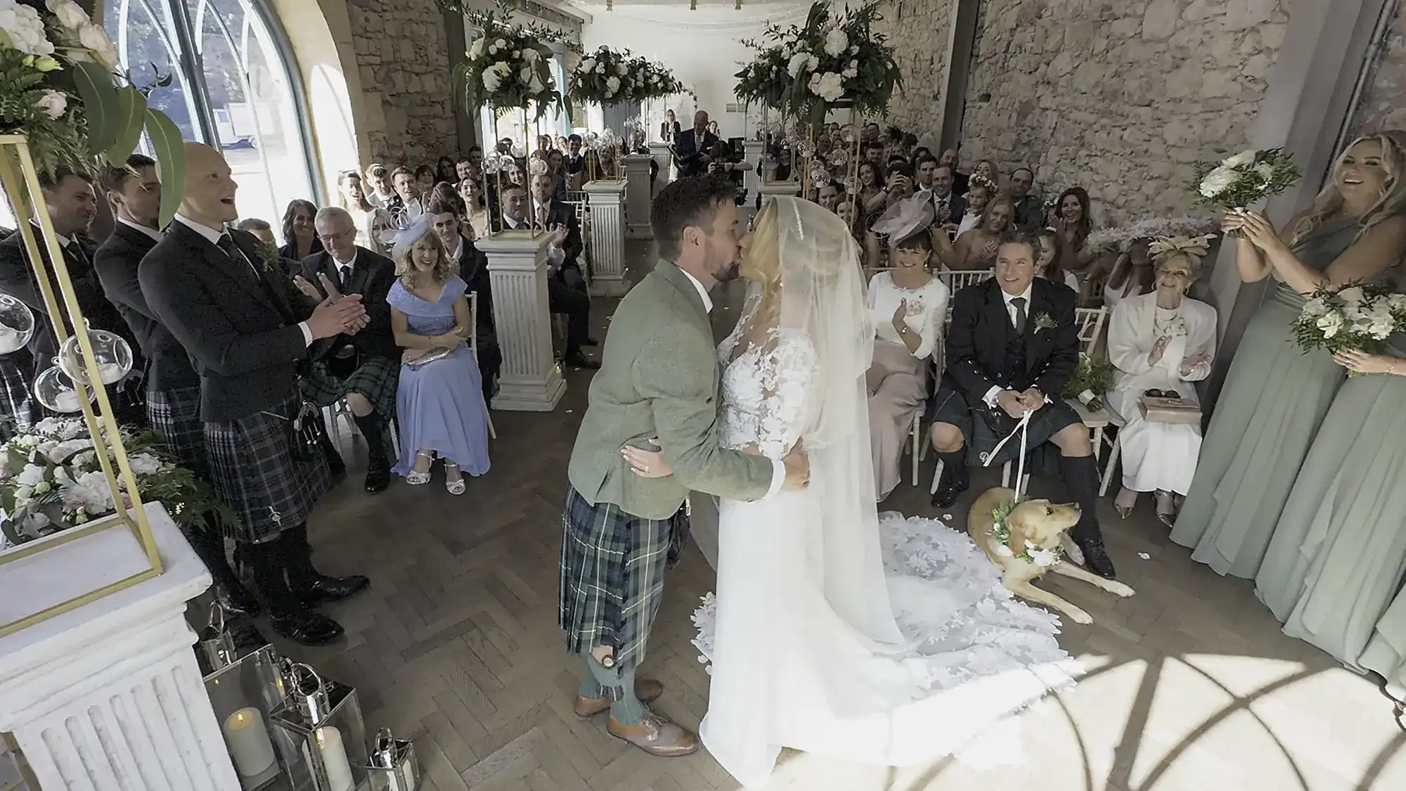 A bride and groom kiss at the altar in a rustic stone venue, surrounded by seated guests. An audience member claps, and a dog lays near the front on the right side.