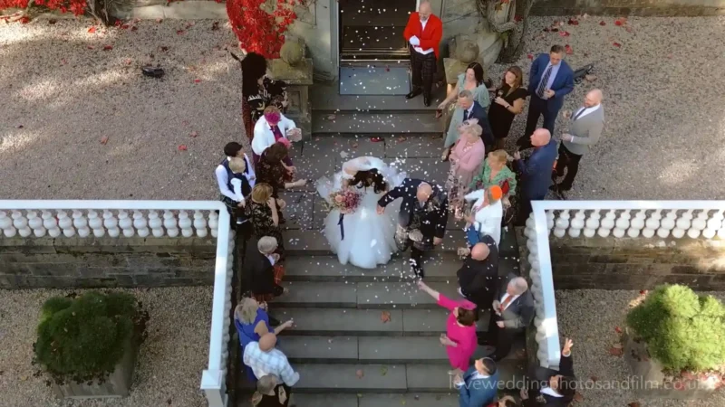 A bride and groom kiss on outdoor stairs as guests surround them, throwing flower petals. The scene is viewed from above.