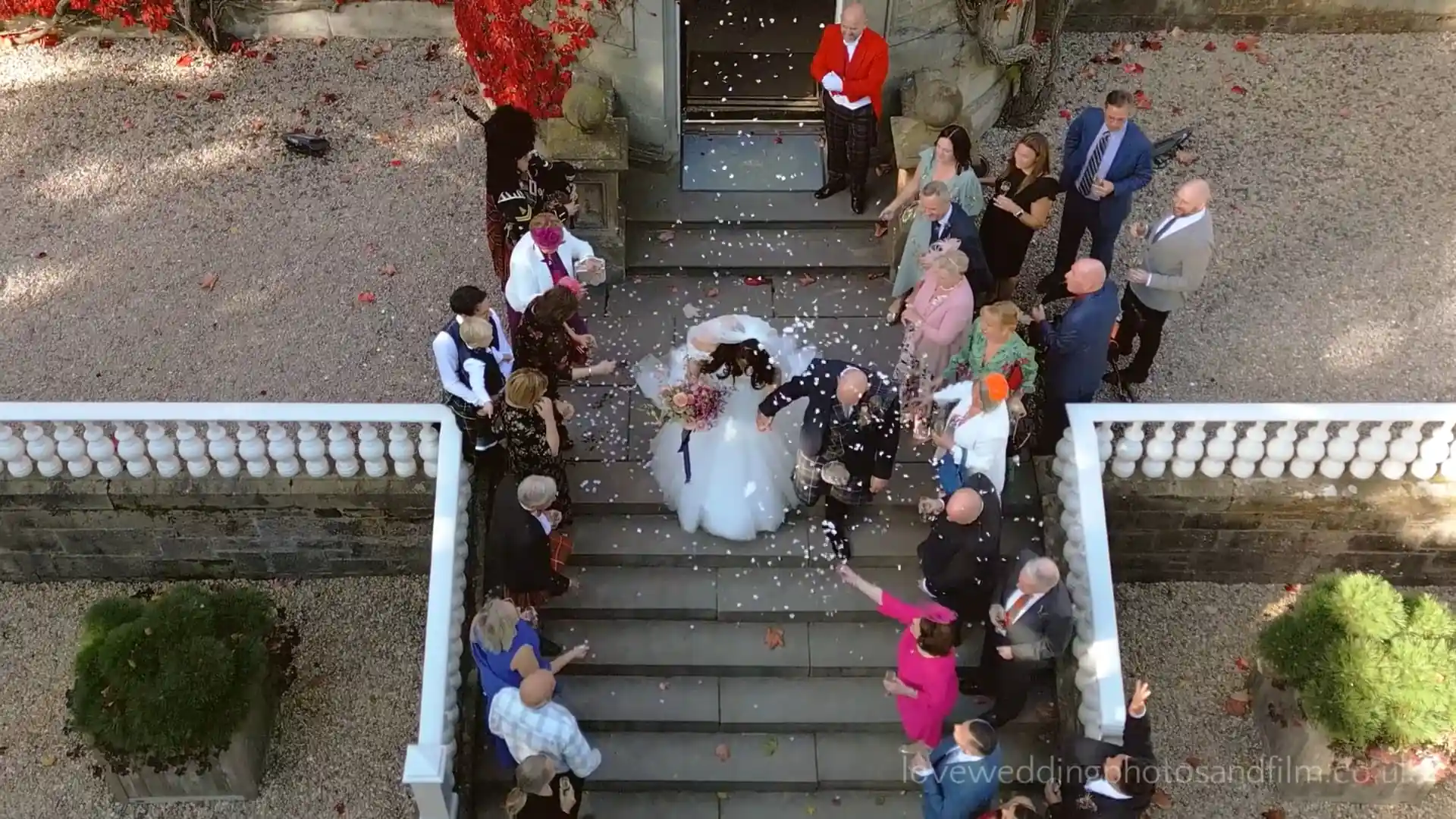 A bride and groom kiss on outdoor stairs as guests surround them, throwing flower petals. The scene is viewed from above.
