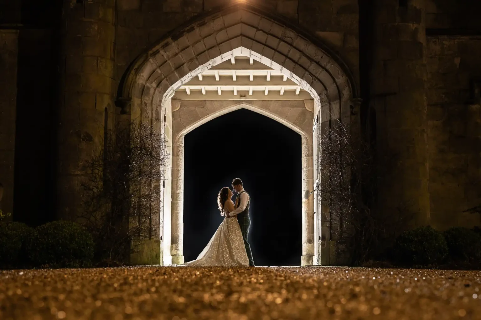 A couple stands embraced under an illuminated Gothic archway at night, with a dark background and a gravel path in the foreground.