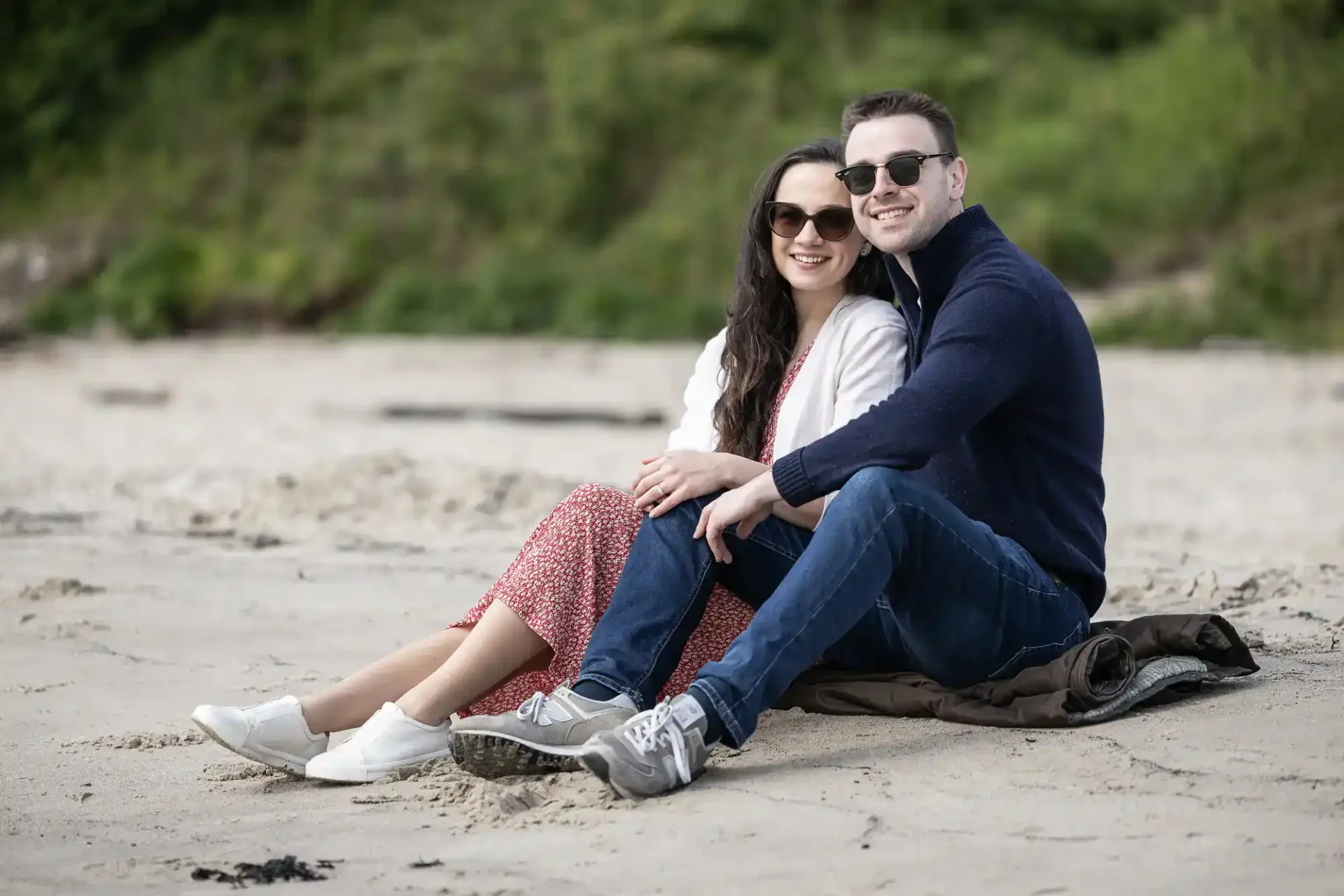 A smiling couple wearing sunglasses and casual clothes sit closely together on a blanket at a sandy Aberdour Beach, with greenery blurred in the background. The woman wears a red dress and white trainers; the man wears a navy jumper, jeans, and grey trainers.