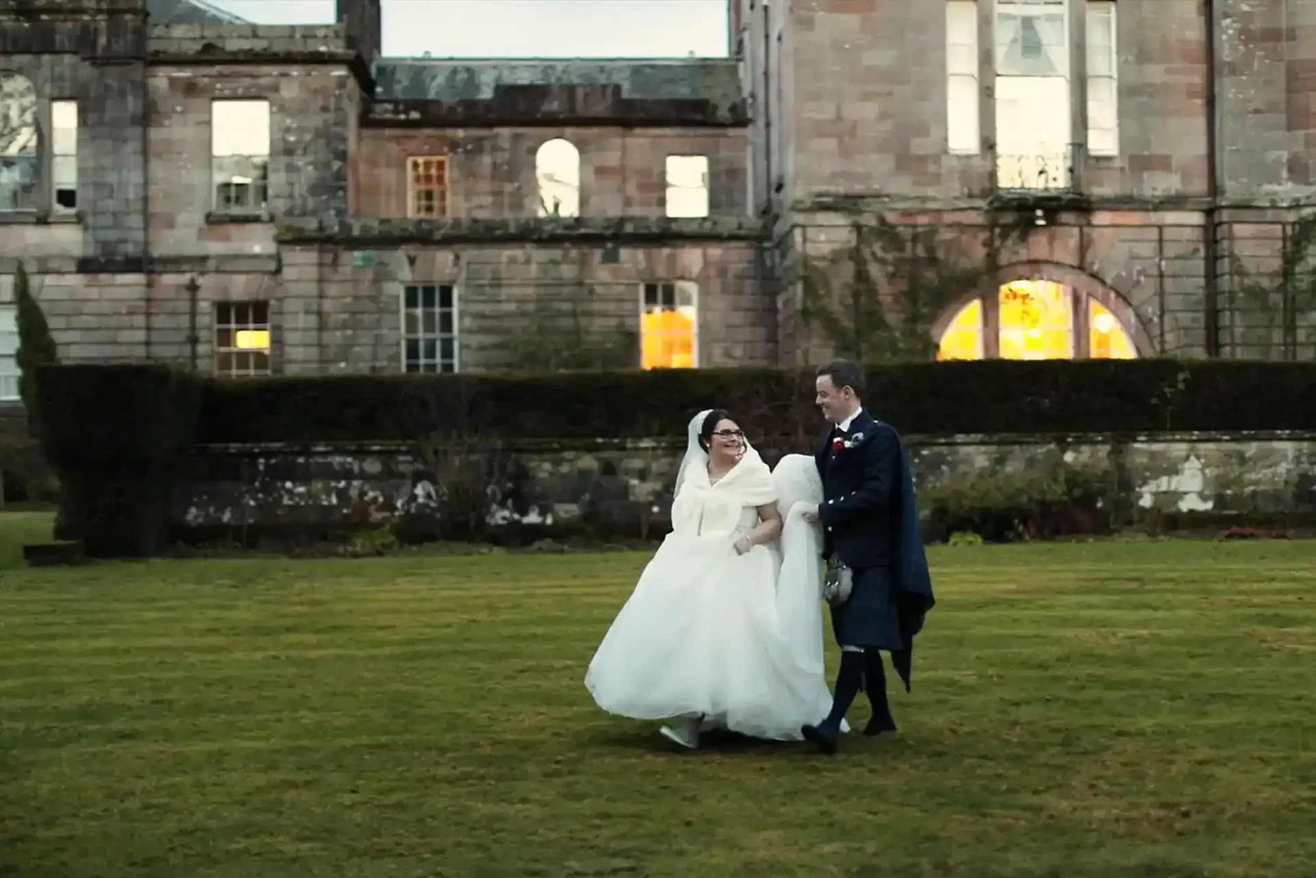 A bride in a white dress and veil walks arm-in-arm with a groom in a dark suit and tartan kilt on the lawn in front of an old stone building with large windows, some lit warmly from inside. Both are smiling, and the scene appears joyful and candid.