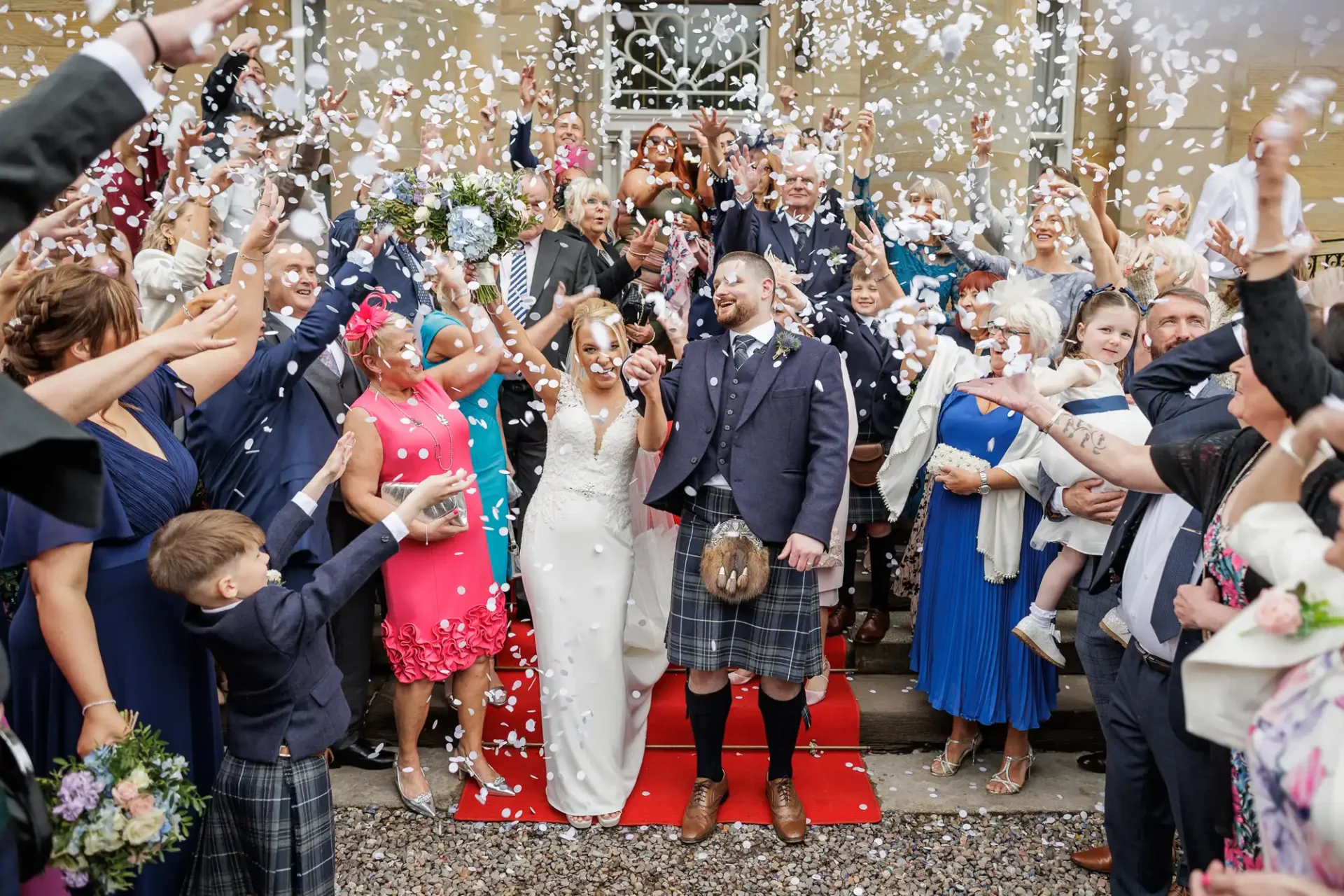 A bride and groom in wedding attire stand on a red carpet surrounded by guests throwing confetti and celebrating outside a building.