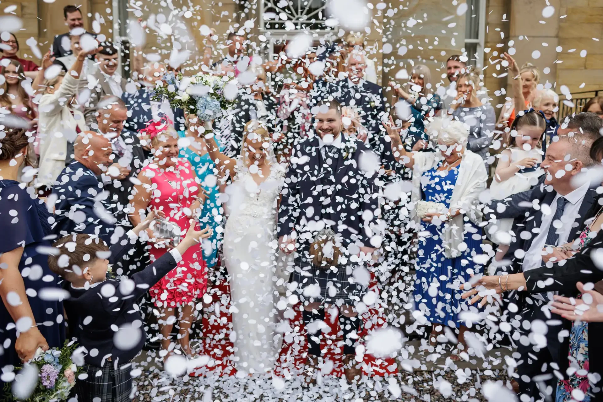 A bride and groom stand surrounded by guests as white confetti falls during a wedding celebration outside a building, filmed by Edinburgh wedding videographer Love Wedding Photos And Film.