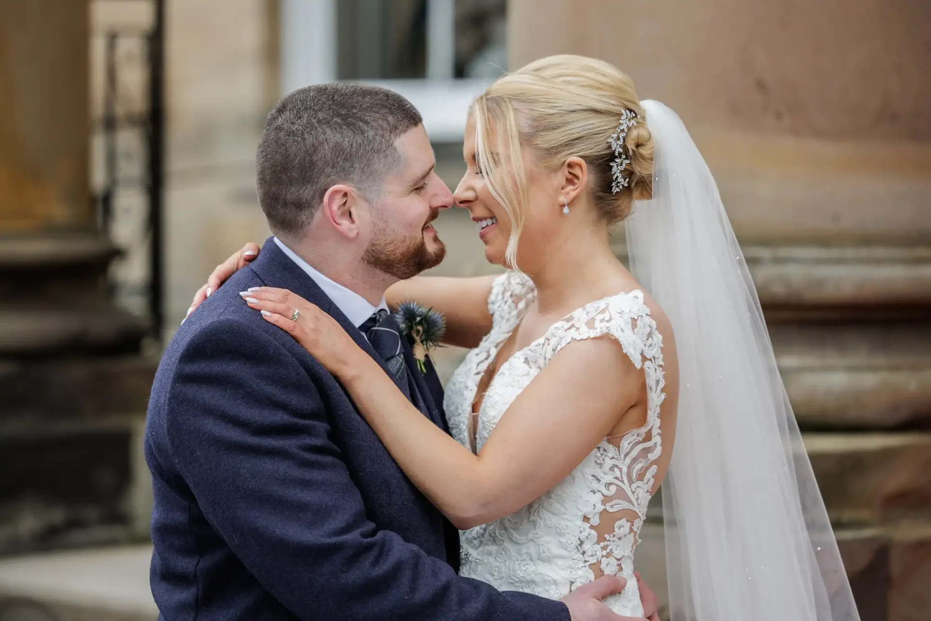 A bride and groom stand close, smiling at each other and embracing outside a stone building. The bride wears a lace dress and veil; the groom wears a suit with a boutonniere.