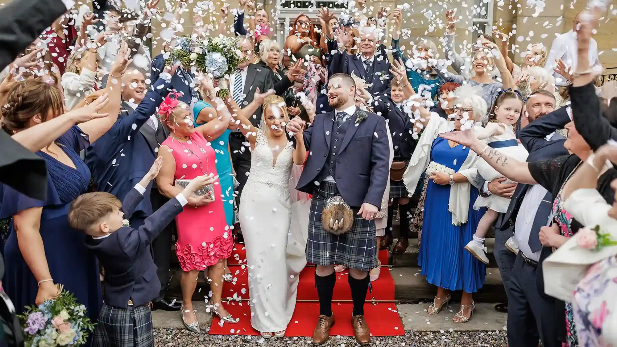 Balbirnie House wedding videographer image of a joyful wedding group stands on steps, tossing white confetti. The bride in a white dress and groom in a kilt are at the centre, smiling. Guests surround them, dressed in colourful attire, celebrating with raised arms and big smiles.