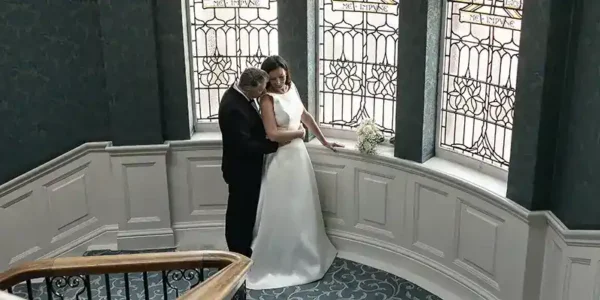 Comprehensive guide to the best Edinburgh wedding venues by local wedding photographer. A bride in a white dress and a groom in a black suit embrace on a curved staircase with ornate patterned carpet. Behind them are tall stained glass windows and white paneled walls. A small bouquet sits on the ledge next to them.