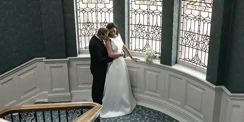 Comprehensive guide to the best Edinburgh wedding venues by local wedding photographer. A bride in a white dress and a groom in a black suit embrace on a curved staircase with ornate patterned carpet. Behind them are tall stained glass windows and white paneled walls. A small bouquet sits on the ledge next to them.