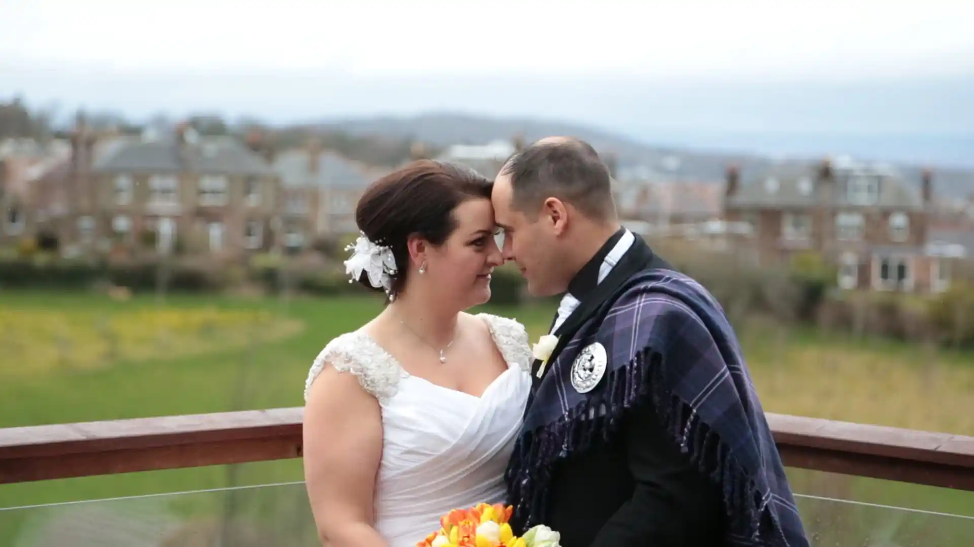 A bride in a white dress and a groom in a dark suit with a tartan sash stand close together on a balcony, touching foreheads and smiling, holding a bouquet. The background shows green grass, houses, and a cloudy sky.