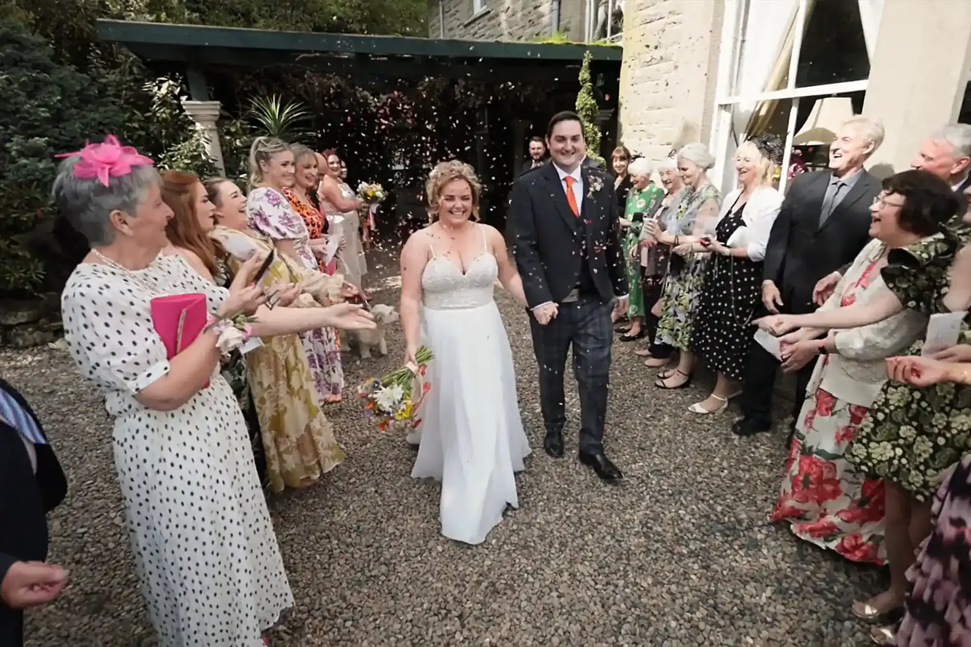 A bride in a white dress and groom in a dark suit walk hand-in-hand on a gravel path at Branxholm Park, smiling, as guests in colourful dresses and suits stand on both sides, celebrating and tossing confetti outside a stone building with greenery in the background.