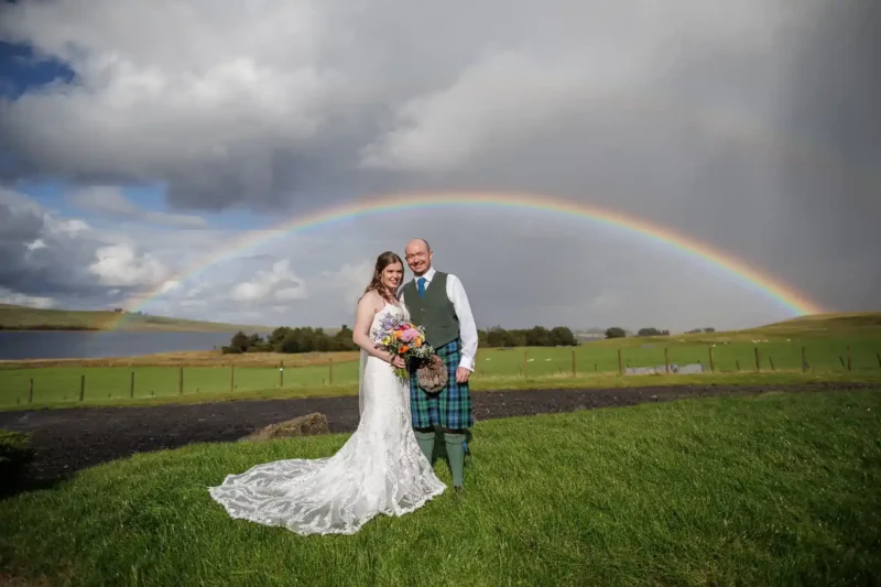 A bride and groom pose outdoors on grass, with a lake, hills, and a rainbow in the background. at Cairns Farm. The bride wears a white dress and holds a bouquet; the groom wears a kilt. West Lothian wedding venues page.