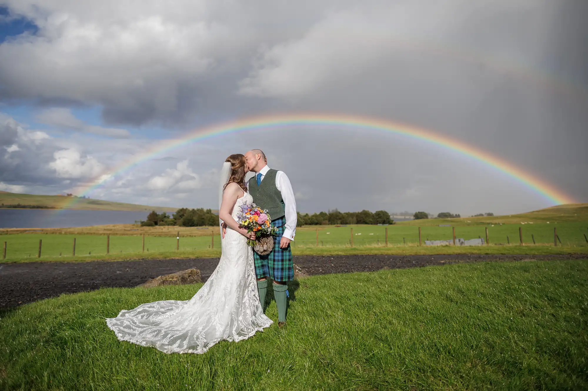 Cairns Farm wedding photographer photo of a bride in a white lace dress and a groom in a tartan kilt kiss outdoors on green grass, under a vibrant rainbow stretching across a cloudy sky. The rural landscape features a lake and distant trees, creating a romantic and scenic backdrop.