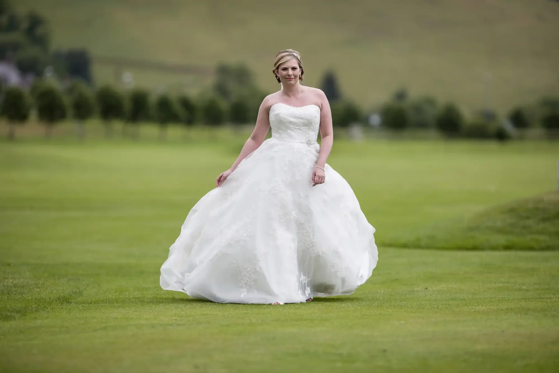 A bride in a white strapless wedding dress stands on a green grassy golf course at Cardrona Hotel Peebles. She is holding the sides of her flowing dress and smiling softly. The background features blurred trees and rolling hills under a cloudy sky.