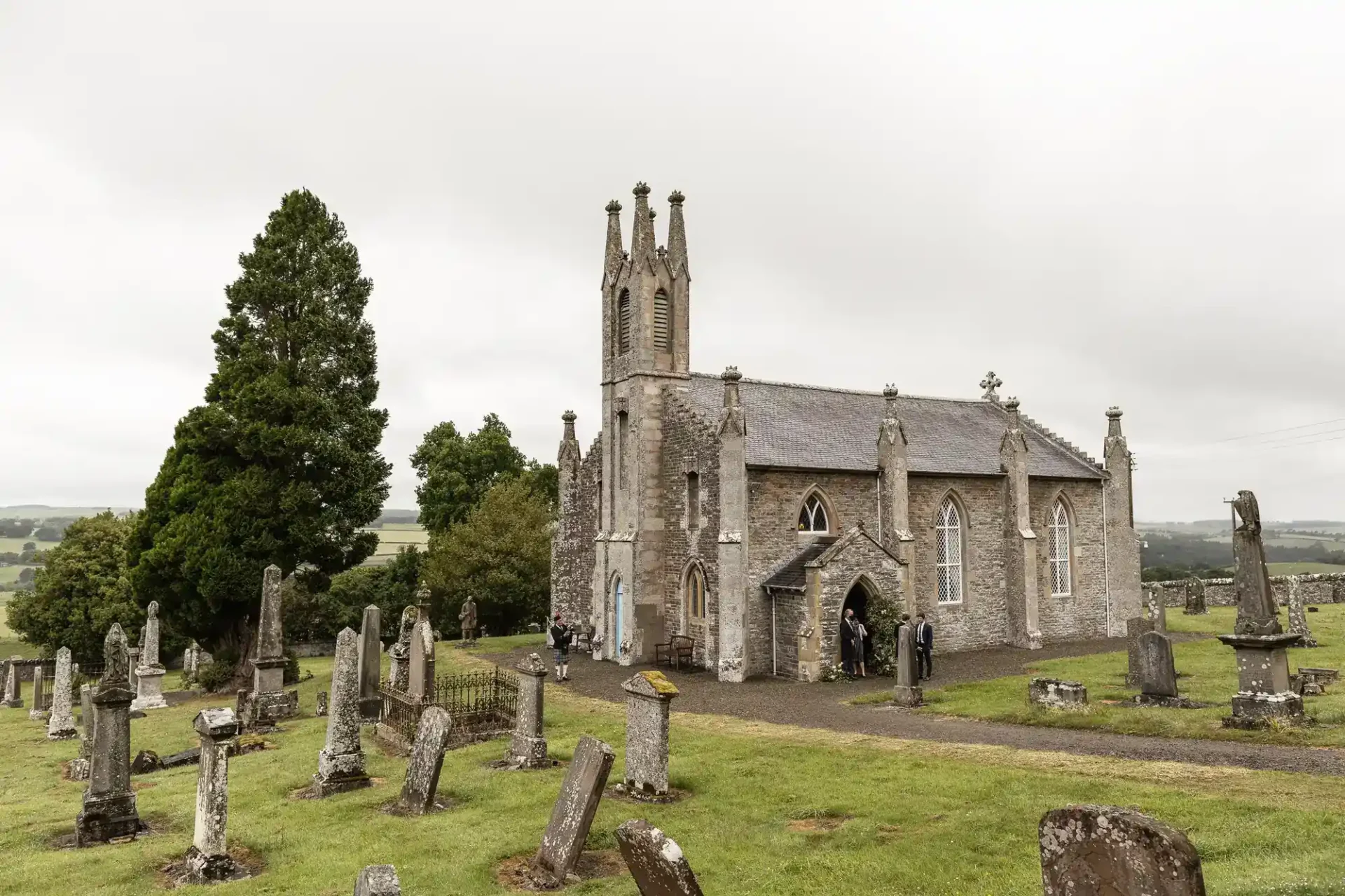Cavers & Kirkton Parish Church, with pointed arches and a tall central tower stands amid a cemetery of old, weathered gravestones on a grassy hill. People gather near the entrance. Large trees and rolling countryside are visible under an overcast sky.
