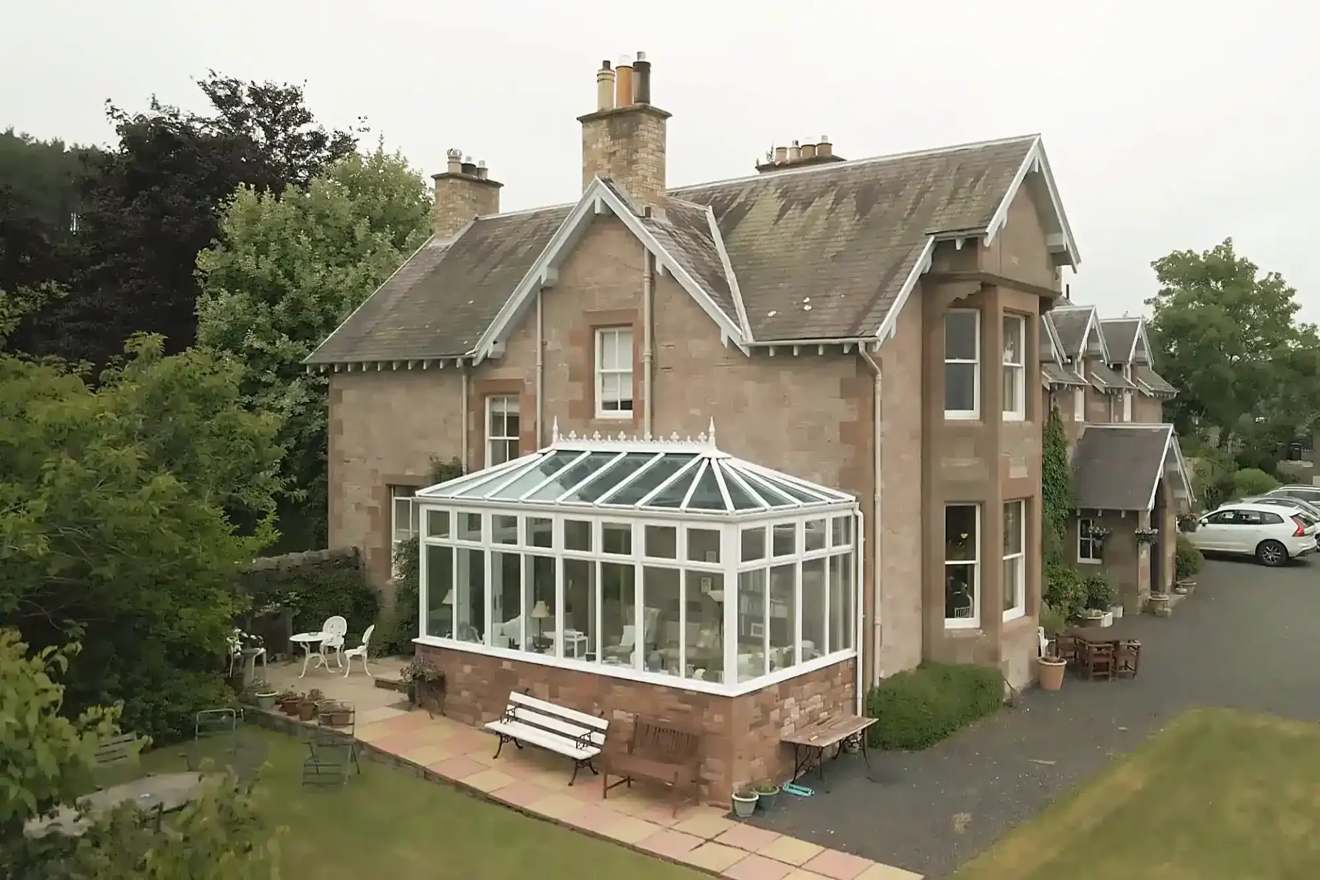 A large, two-storey stone house, Clint Lodge, with a gabled roof and chimneys. Attached is a spacious glass conservatory with a sloping roof. The patio has outdoor furniture, a bench, and is surrounded by green lawn, trees, and a small gravel drive with parked cars.