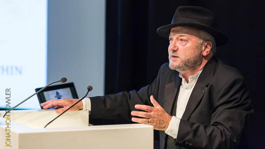 A man in a black hat speaking at a podium with a laptop and a projection screen displaying text behind him.