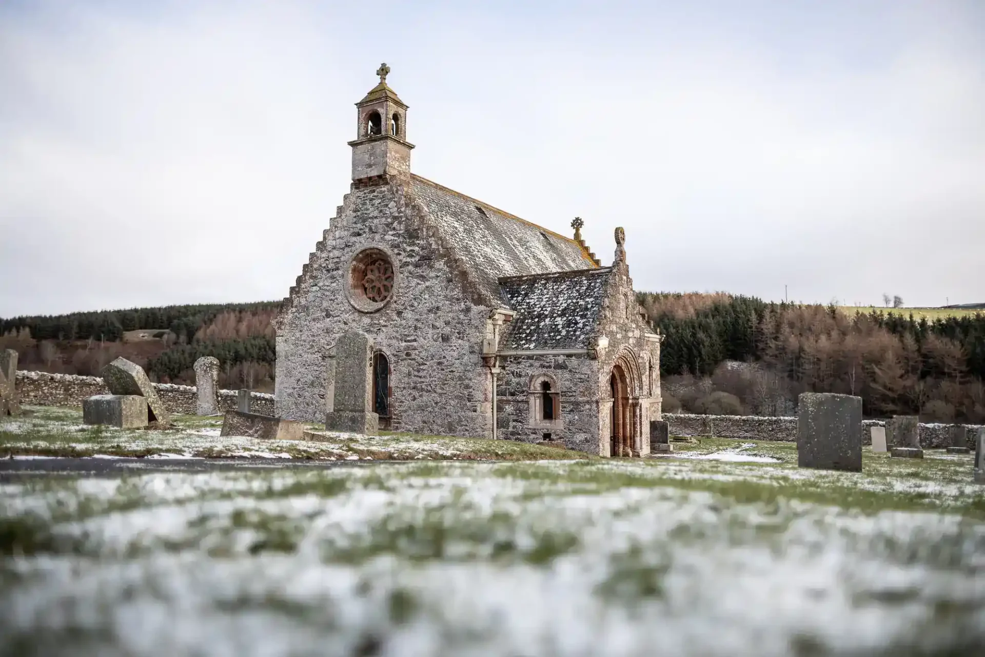 Cranshaws Village Church with a bell tower stands in a snow-dusted graveyard. The church has a circular stained-glass window and arched doorway. Weathered headstones surround the building. Leafless trees and rolling hills are seen in the background.