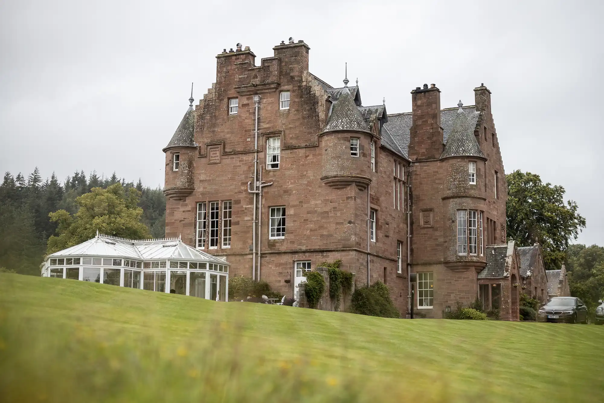 A large, historic stone mansion with turrets and pitched roofs sits on a green lawn. Attached to the left side is a glass conservatory. The sky is overcast, and trees are visible in the background.