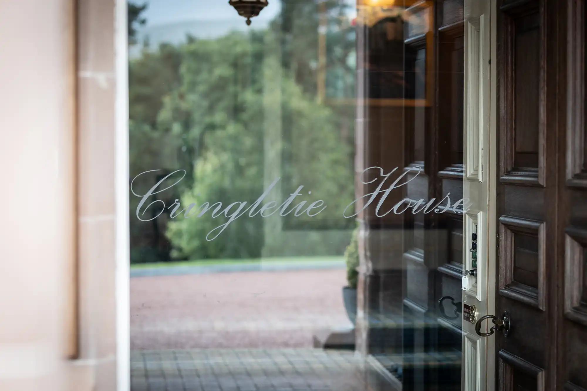 A close-up of a glass door with the words “Cringletie House” elegantly etched in cursive. The wooden door is open, revealing a reflection of trees and outdoor paving, with soft, natural light illuminating the scene.