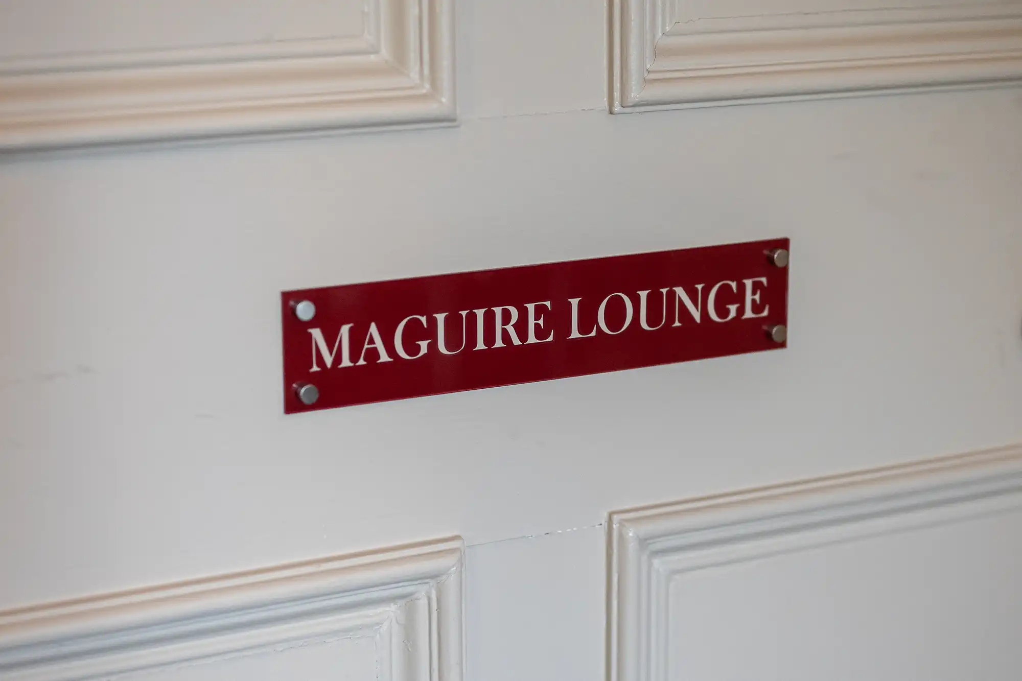 A white panelled door with a red rectangular plaque that reads "MAGUIRE LOUNGE" in bold white capital letters. The sign is attached to the door with four visible screws, one in each corner.
