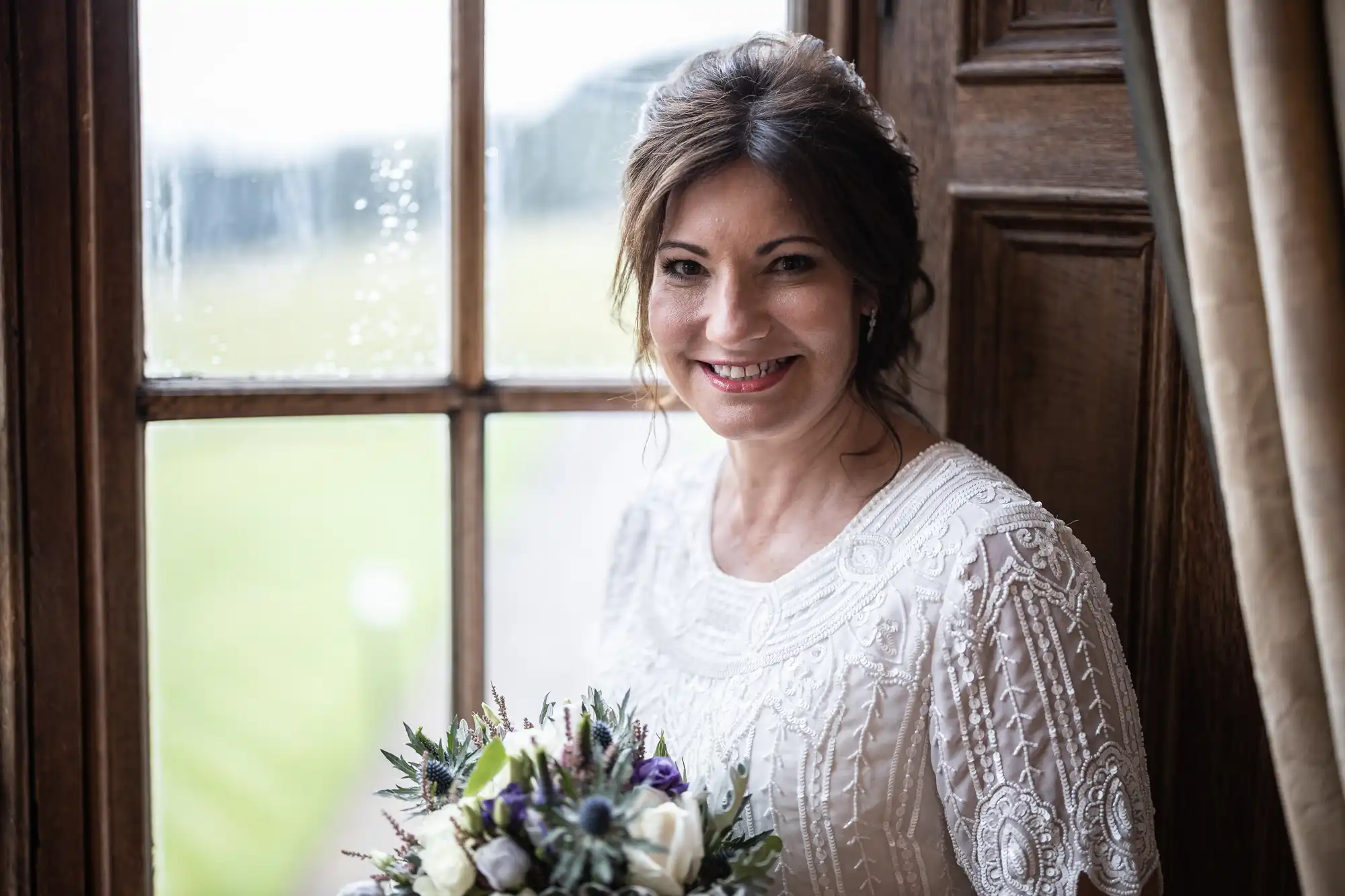 A woman in a white, intricately patterned dress smiles whilst holding a bouquet of white and purple flowers. She stands beside a wooden-framed window with raindrops and beige curtains, with greenery visible outside.