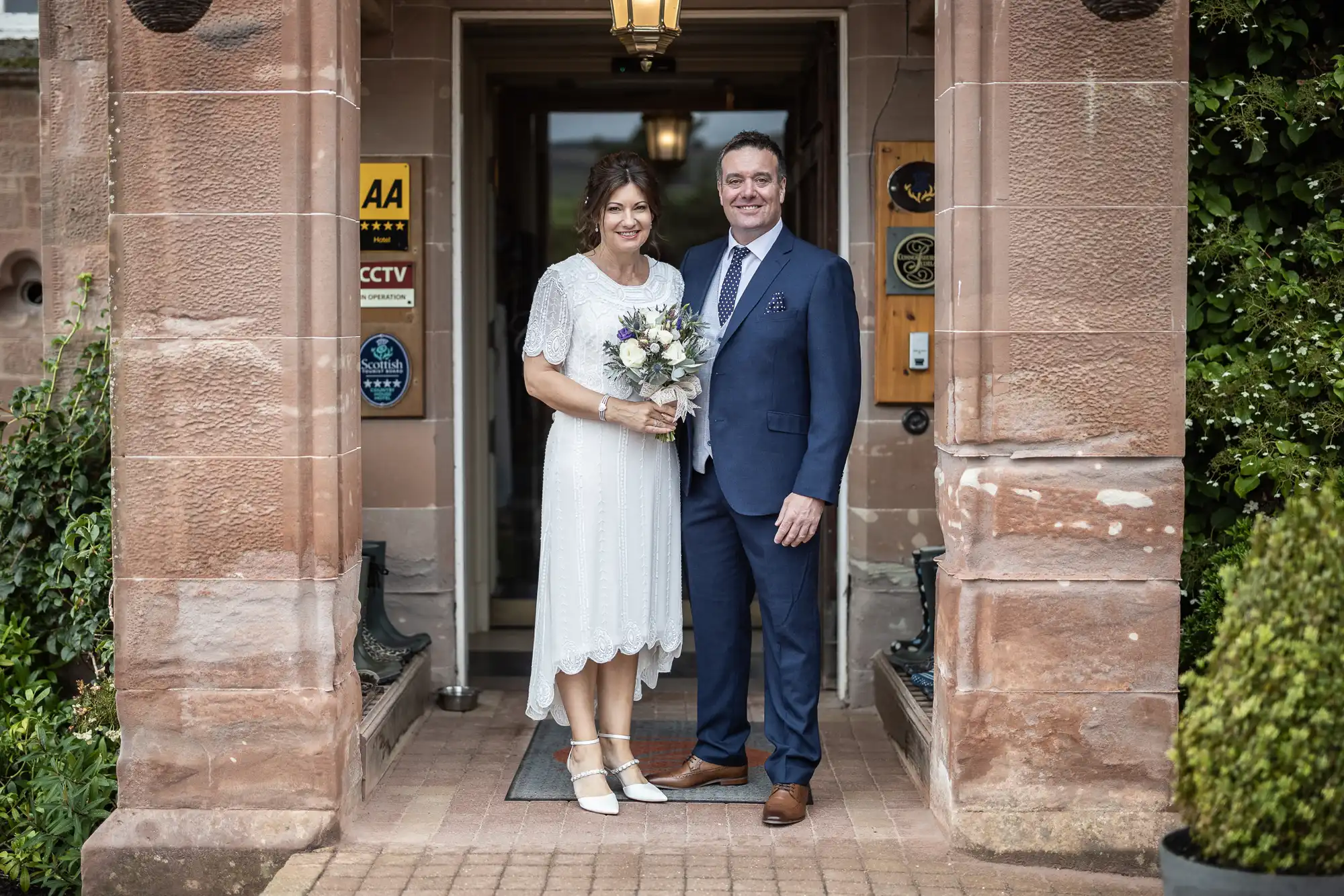 A woman in a white dress holding a bouquet stands beside a man in a blue suit and tie. They smile at the camera, standing in the entrance of a stone building adorned with plaques and plants. The moment appears to be from a formal or celebratory event.